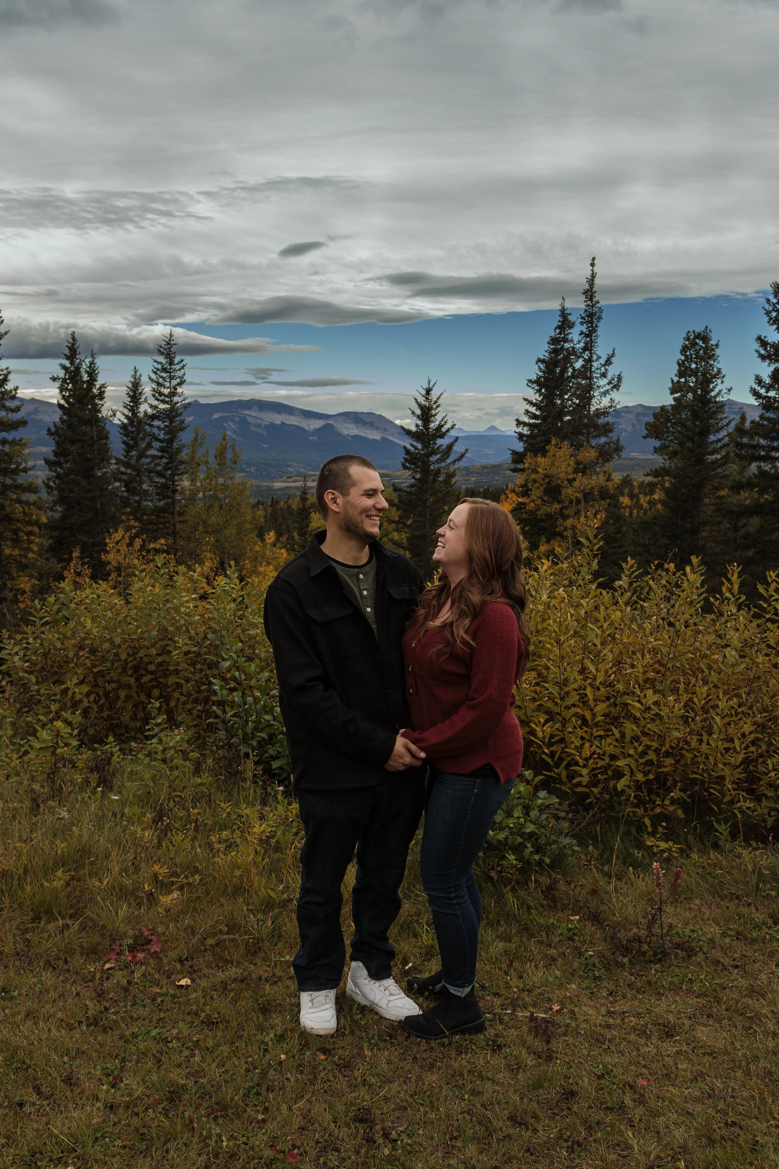 A couple smiling and looking at each other outdoors in a forested area with mountains in the background, under a partly cloudy sky. The man has short hair and a beard, wearing a black jacket, and the woman has long red hair, wearing a red sweater.