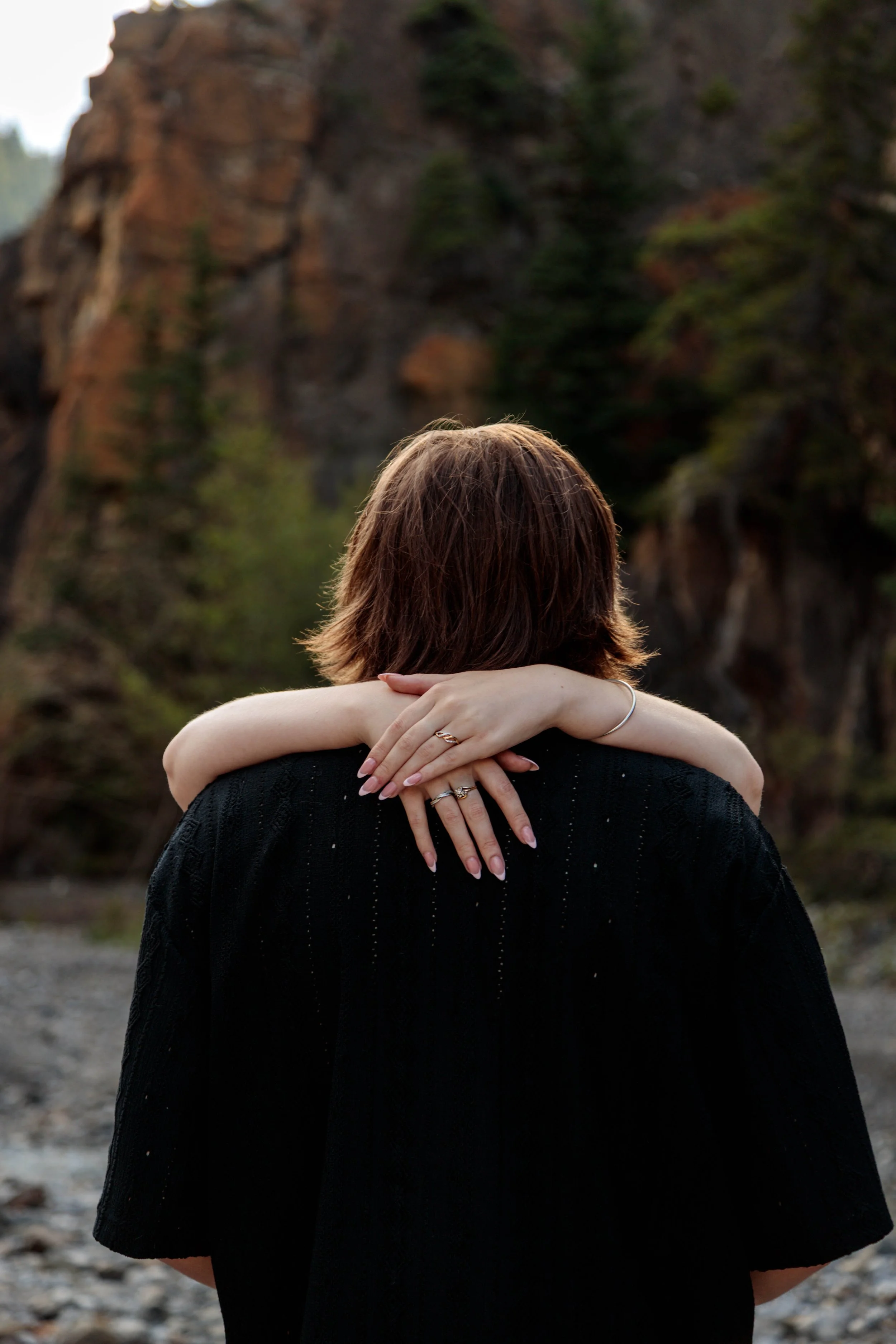 Person with short brown hair in a black sweater, embracing someone with long arms, rings, and a bracelet, outdoors near a rocky, wooded hillside.