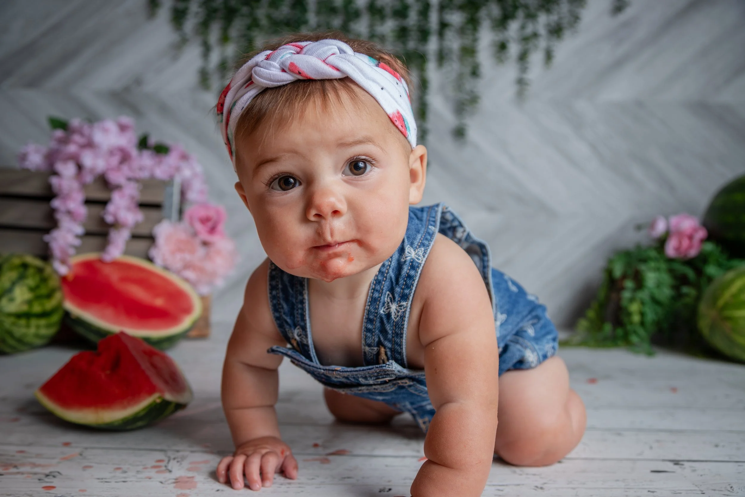 A young child with a headband crawling on a wooden floor surrounded by sliced watermelon and pink flowers.