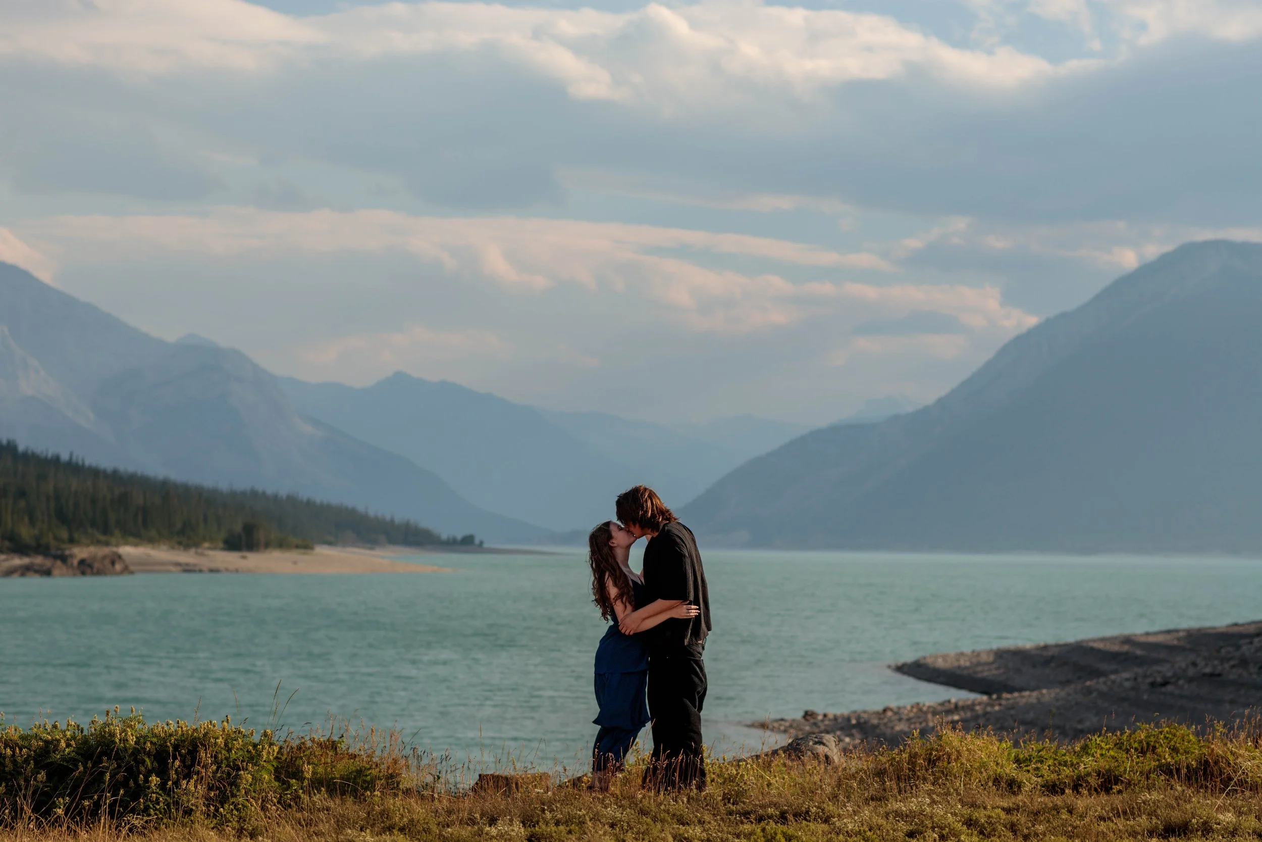 A couple sharing a kiss by Abraham Lake with mountains and beaiutiful sky in the background.