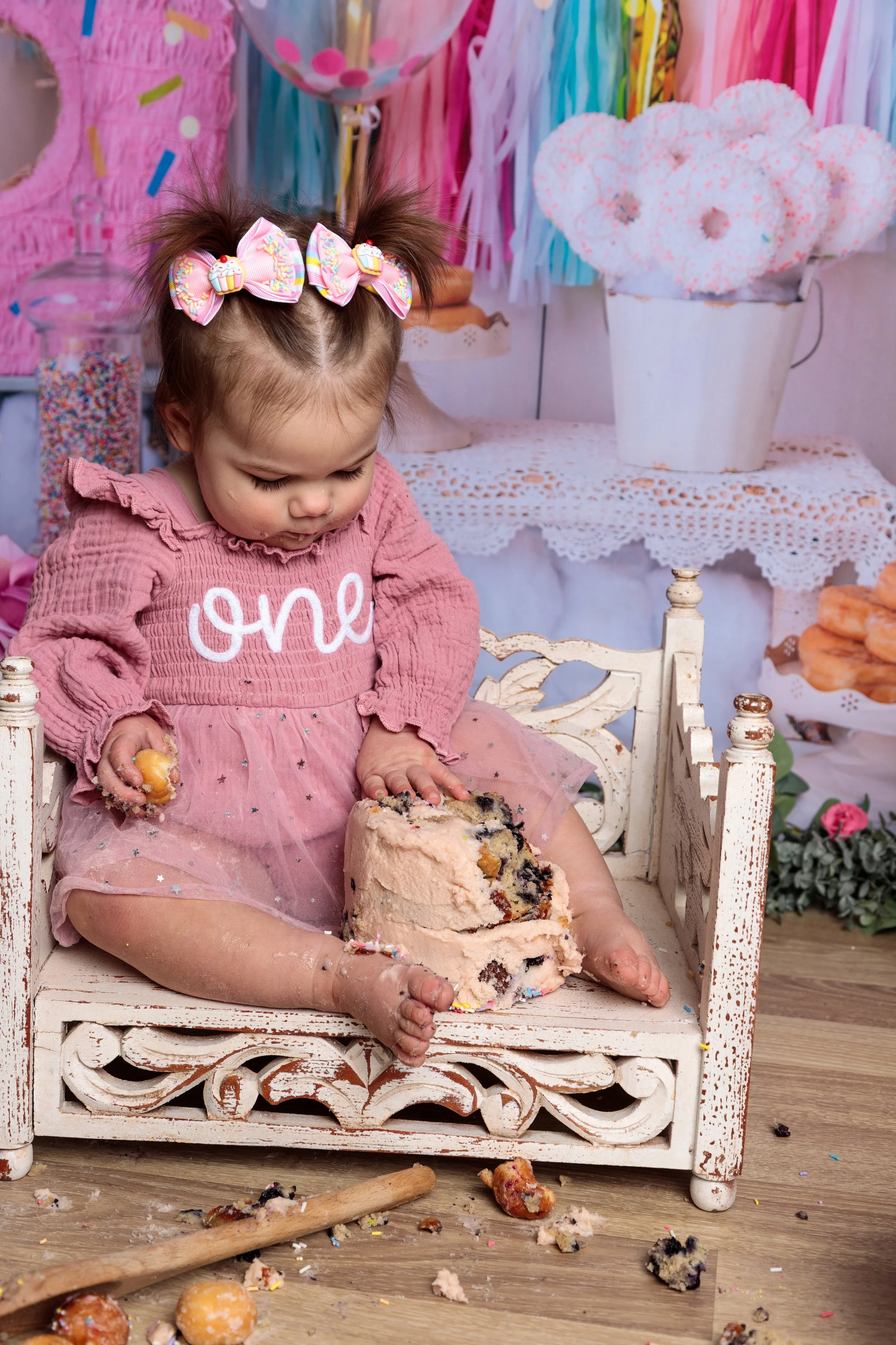 A young girl with messy hands and face, wearing a pink dress with the word 'one' on it, sitting in a small white chair during a cake smash celebration. The girl has a bow with cupcakes on her head and is surrounded by colorful decorations, including 