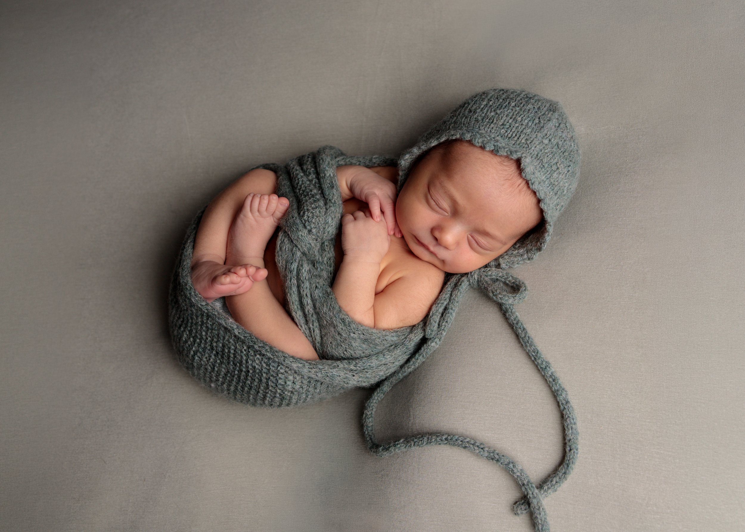 A sleeping newborn baby wrapped in a gray knitted blanket, wearing a matching gray knit hat, lying on a gray surface.