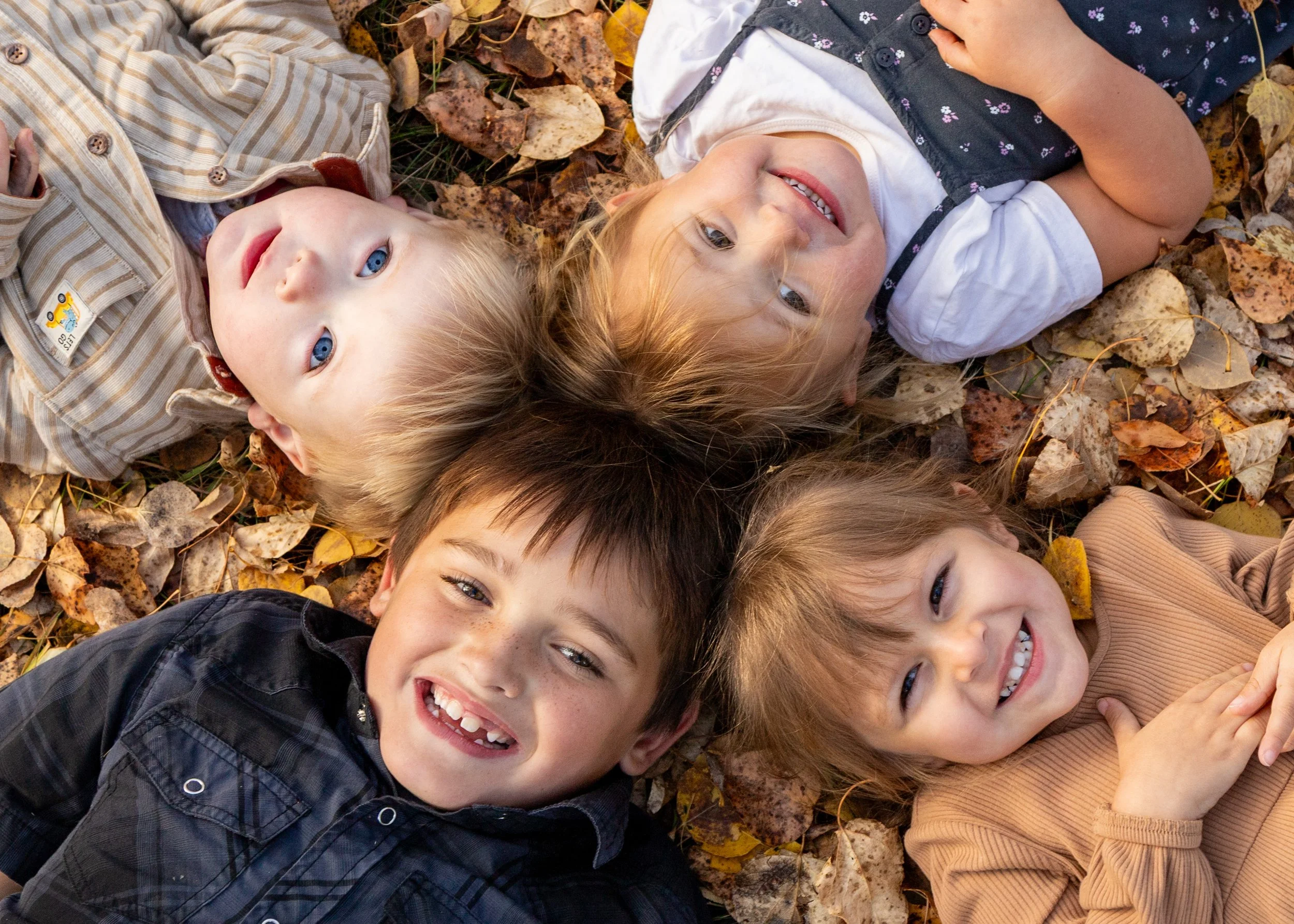 Children lying on the ground covered with autumn leaves, smiling and looking up at the camera.