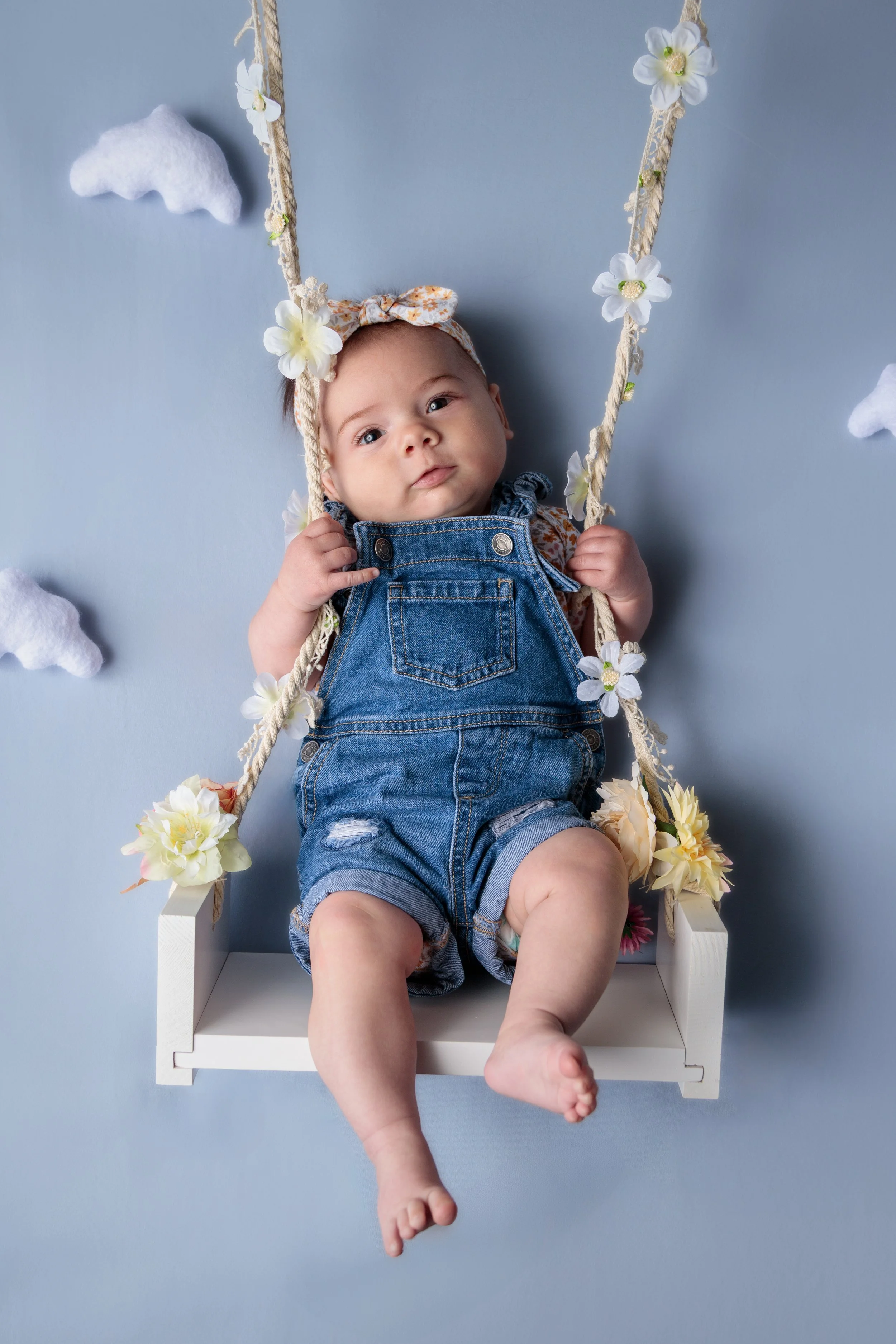 A baby sitting on a swing decorated with white and yellow flowers, against a light blue background. The baby wears denim overalls and a floral headband, looking at the camera.