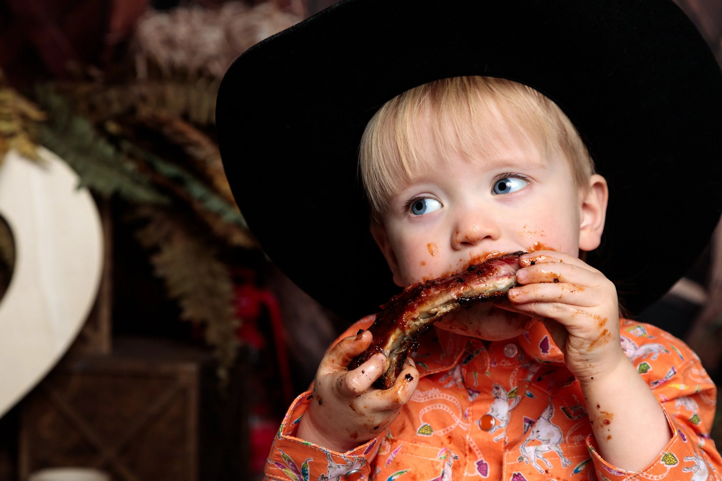 A young child with blond hair and blue eyes eating a large rack of ribs, covered in barbecue sauce. The child is wearing an orange shirt with colorful patterns and a black hat, with barbecue sauce on their face and hands.