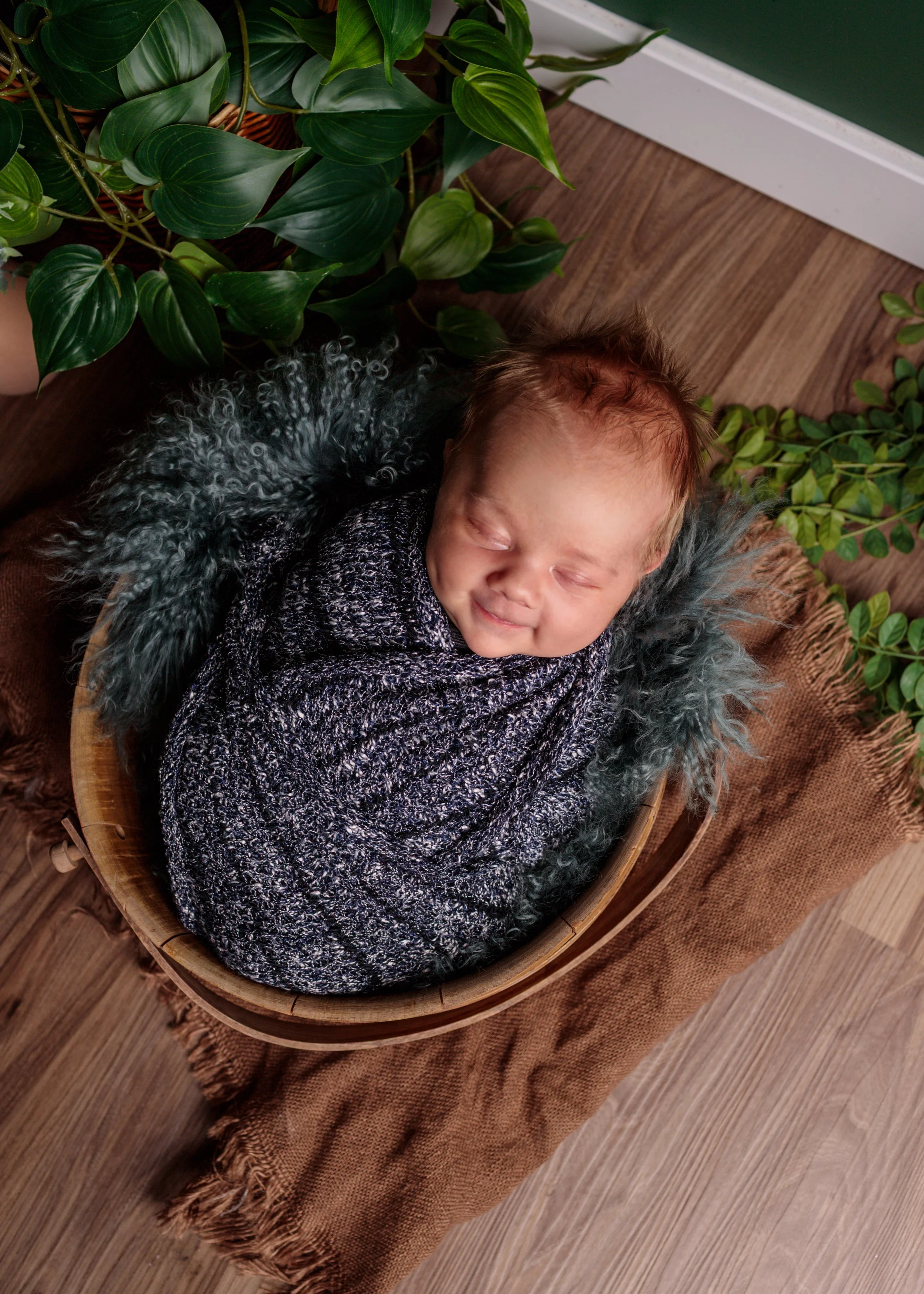 A young child with brown hair sleeping peacefully in a wooden bowl, wrapped in a black and white knitted blanket, surrounded by green plants, on a wooden floor with a burlap cloth underneath.