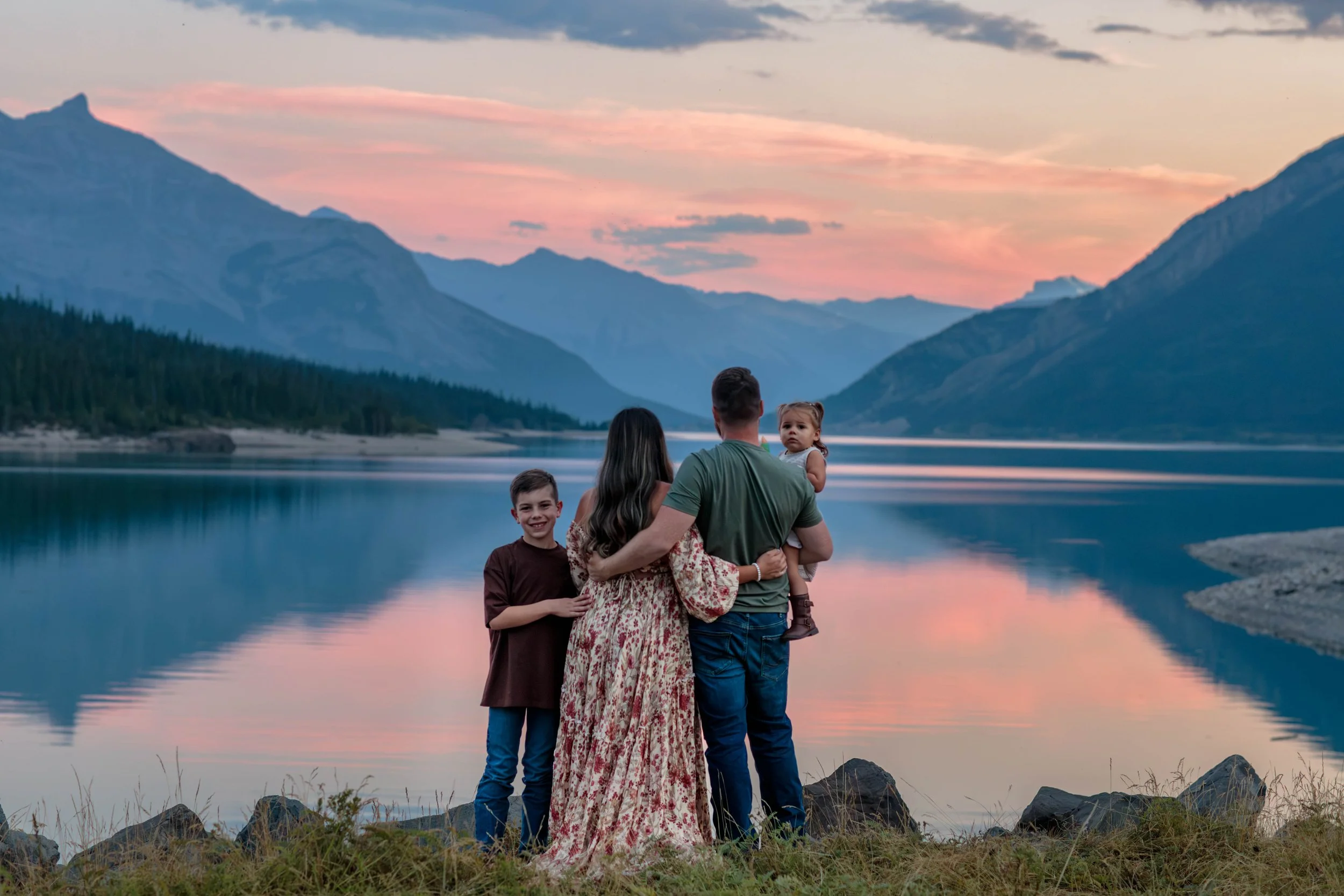 A family of five stands near a lake at sunset, surrounded by mountains and trees.