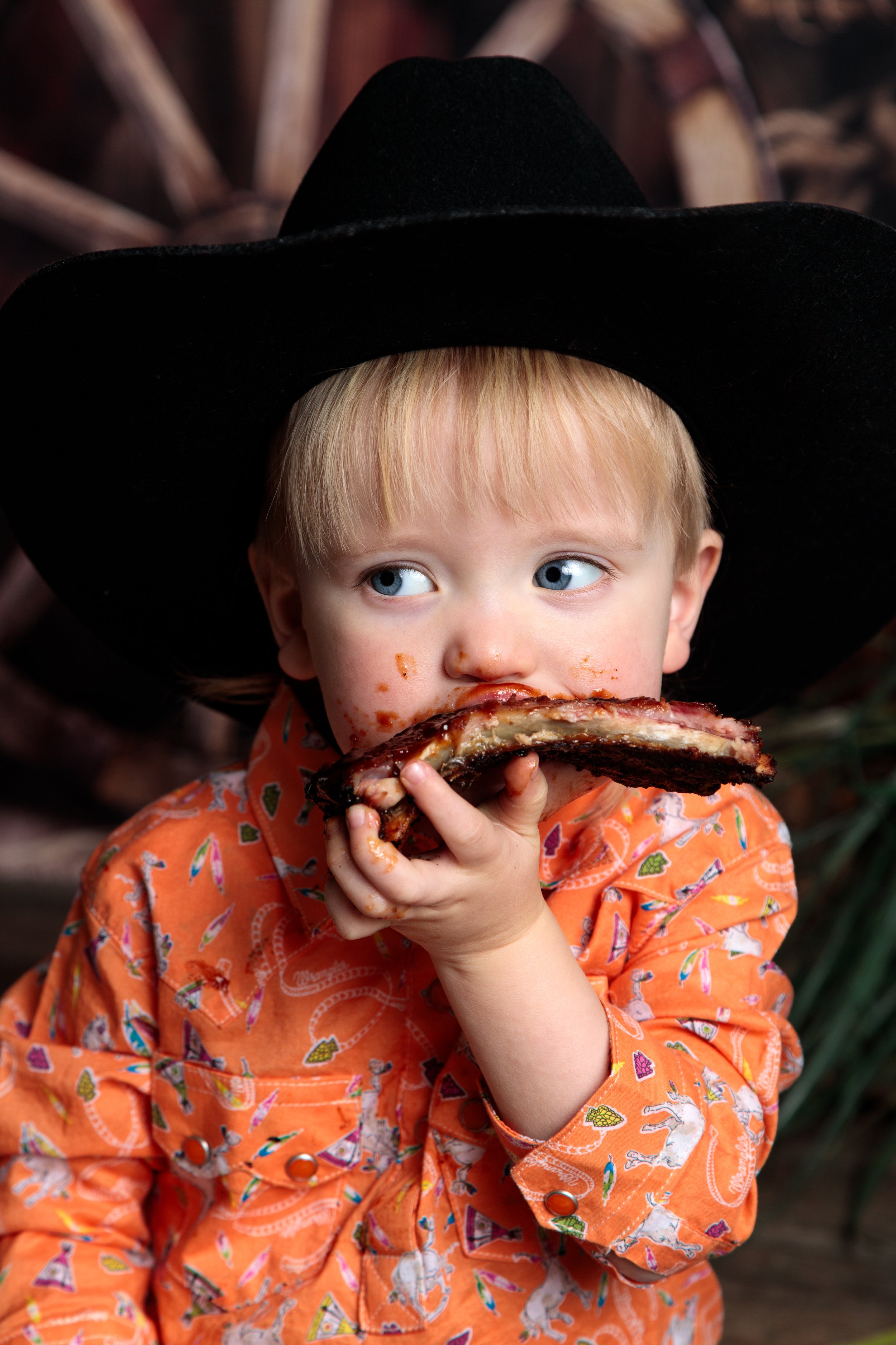 A young child with blonde hair and blue eyes, wearing an orange shirt with colorful patterns, is eating a large piece of barbecue ribs and has food on their face and hands. The child is wearing a large black hat and is outdoors with a rustic wooden b