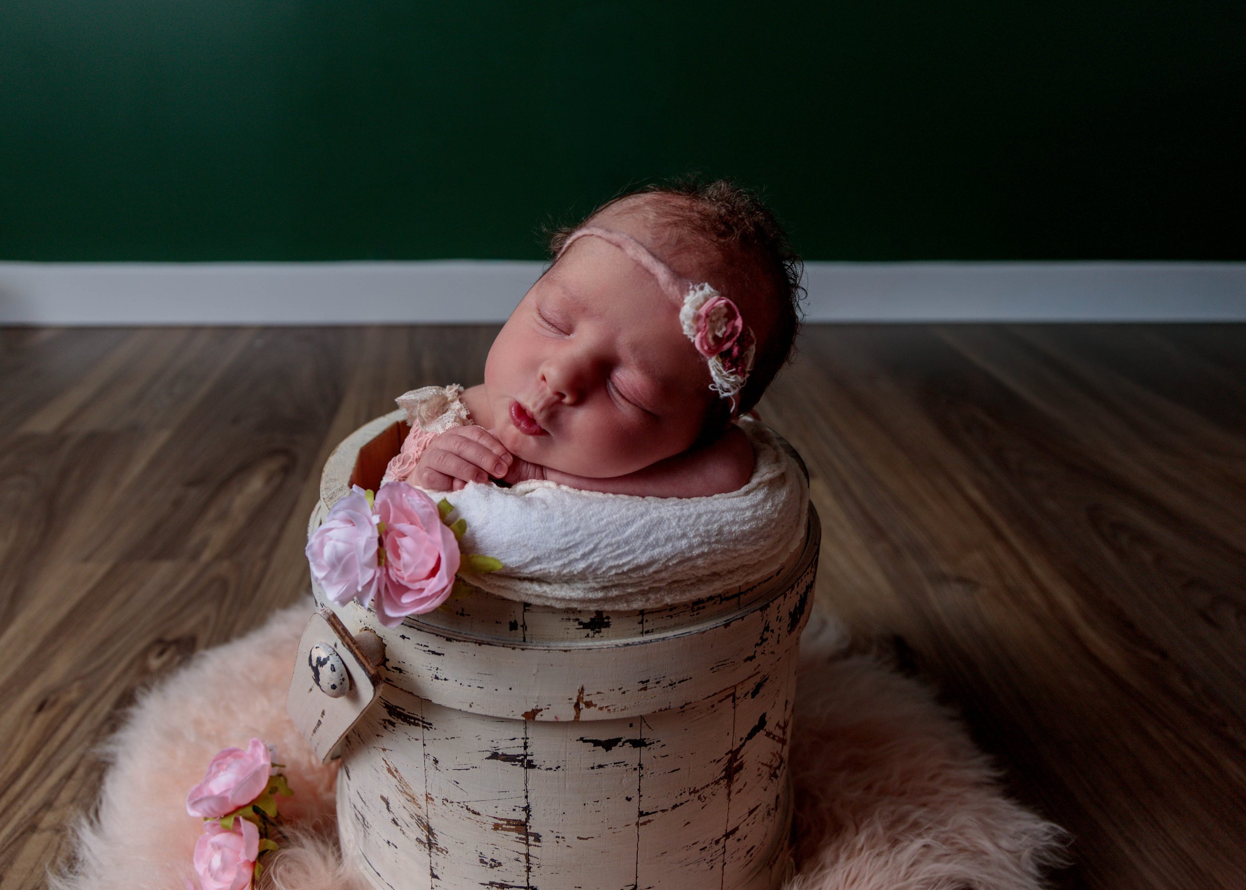 A sleeping baby with a headband, placed in a rustic bucket, decorated with pink flowers, sitting on a pink fluffy rug, with a green wall in the background.