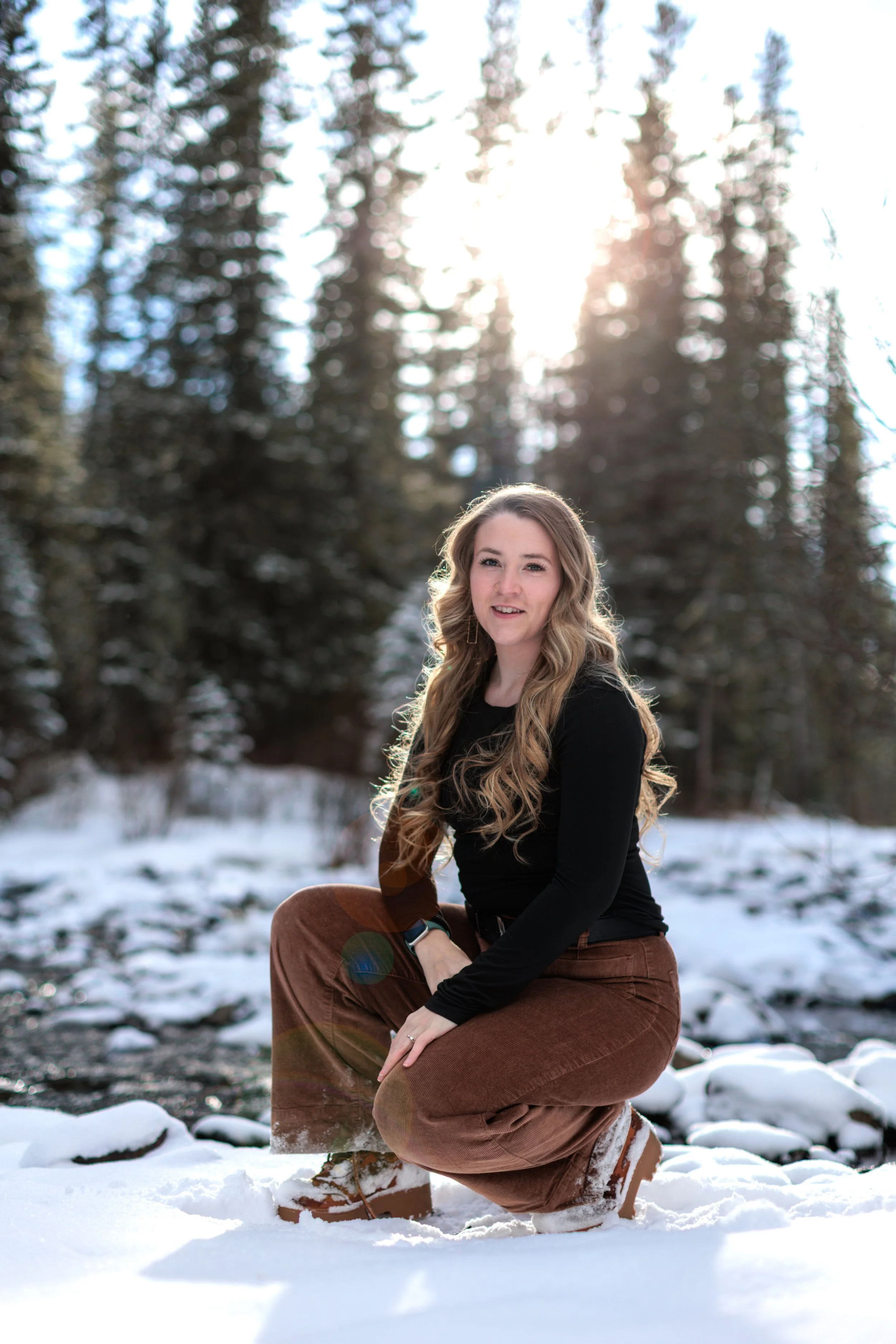A young woman with long wavy hair, wearing a black long-sleeve top and brown pants, kneeling on snow-covered ground in a forest with tall evergreen trees, backlit by the sun.