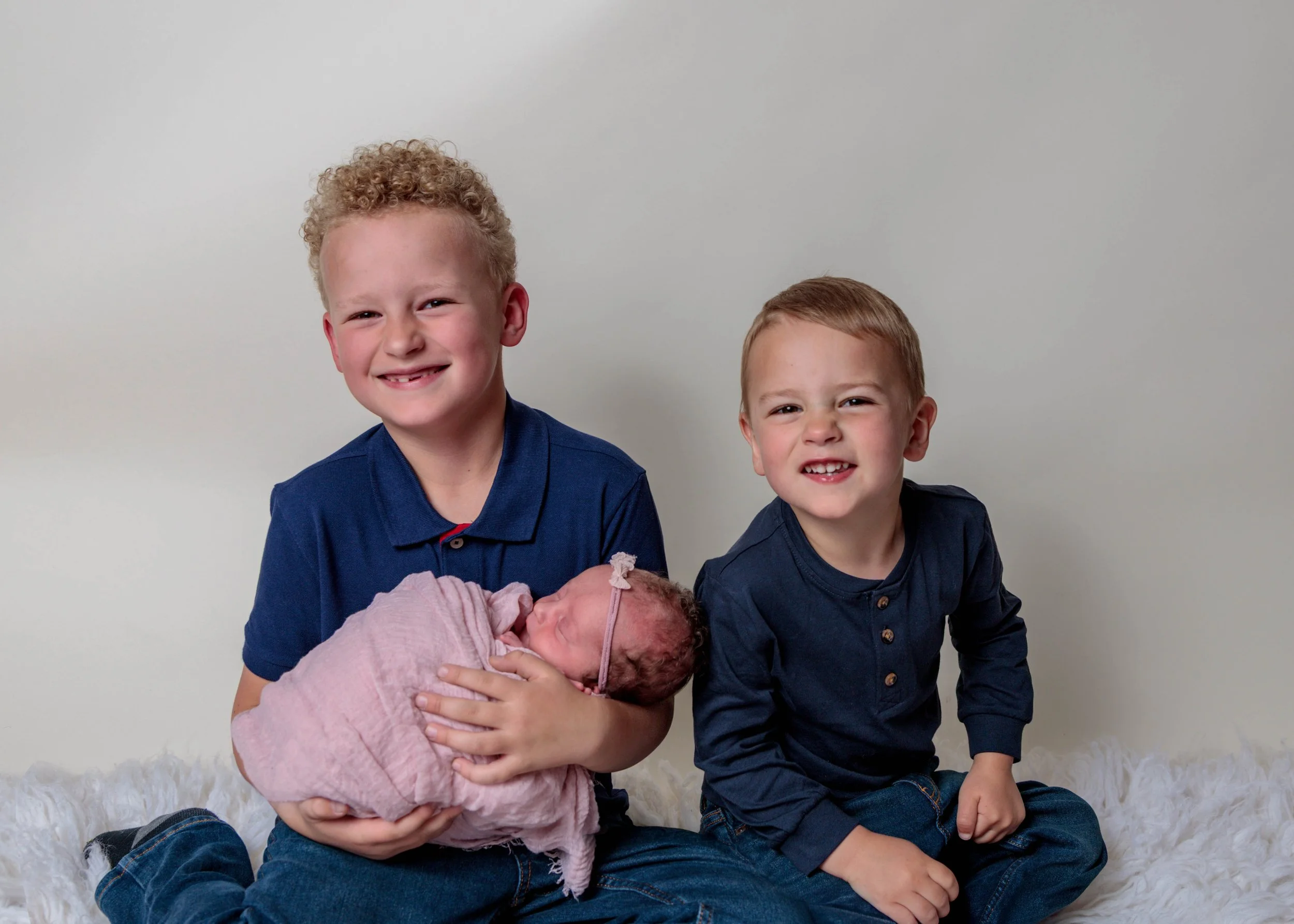 Two young boys, one with curly blond hair and the other with straight red hair, holding a newborn baby wrapped in pink blanket, sitting on a white furry surface against a plain white background.