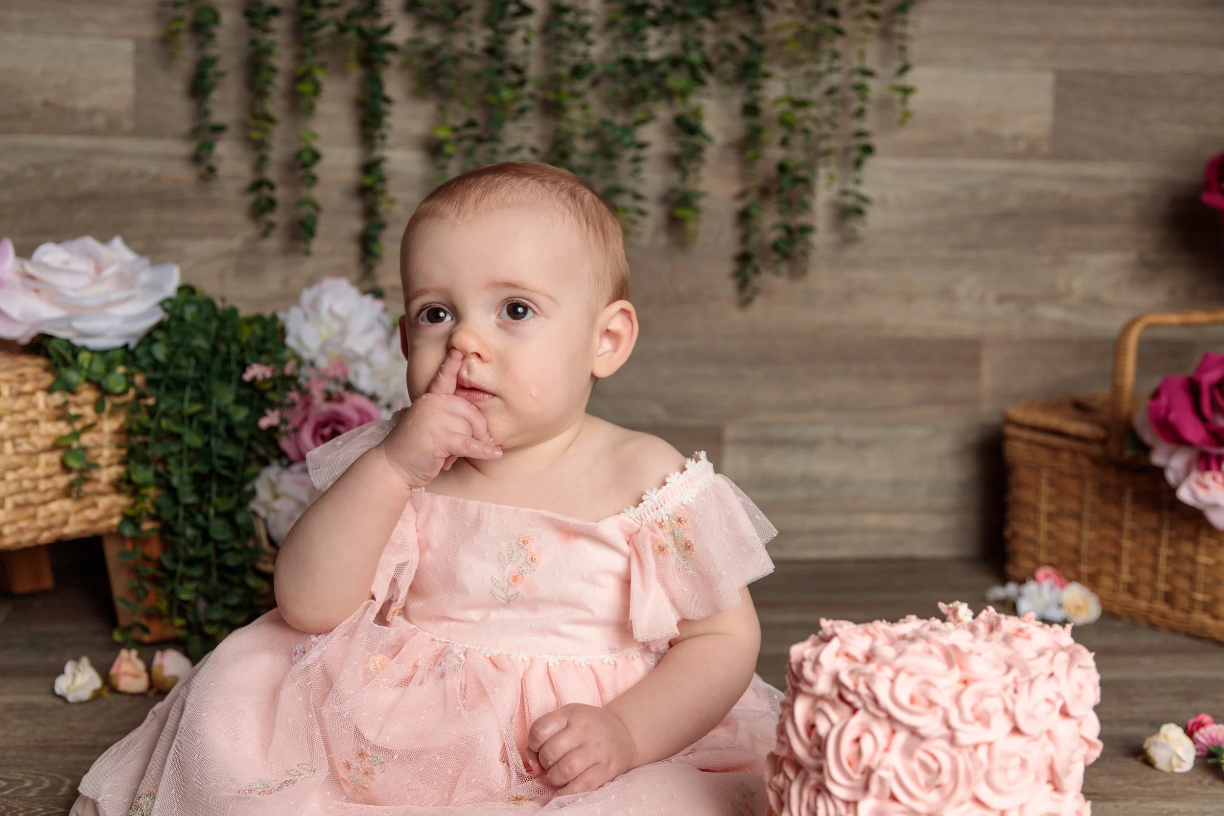 A young girl in a pink dress with embroidered flowers sitting in front of a decorated backdrop with flowers and greenery. She has her finger in her nose and appears to be looking upwards.