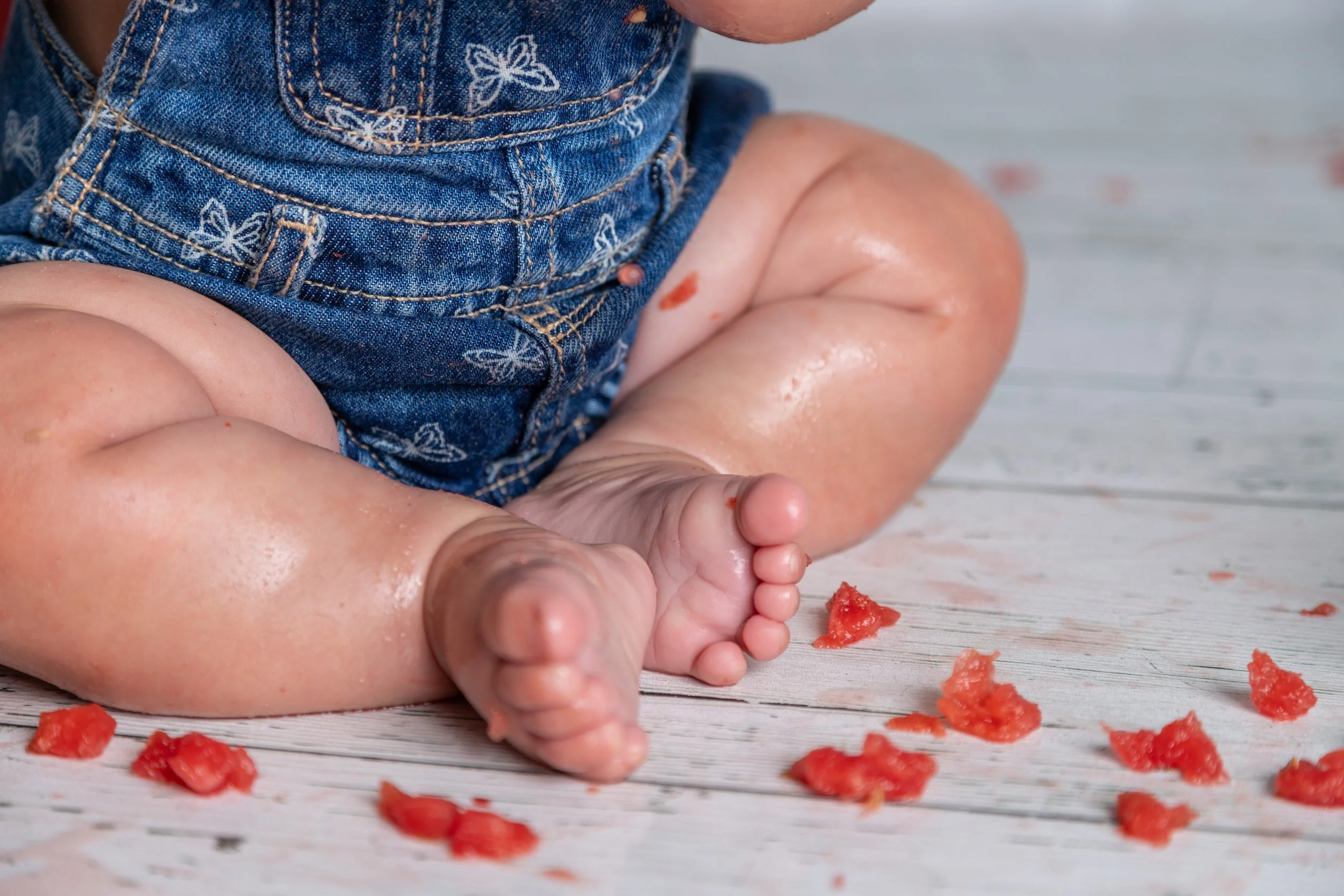 Close-up of a baby crawling on a white wooden floor with scattered pieces of watermelon, wearing a denim outfit with butterfly embroidery.