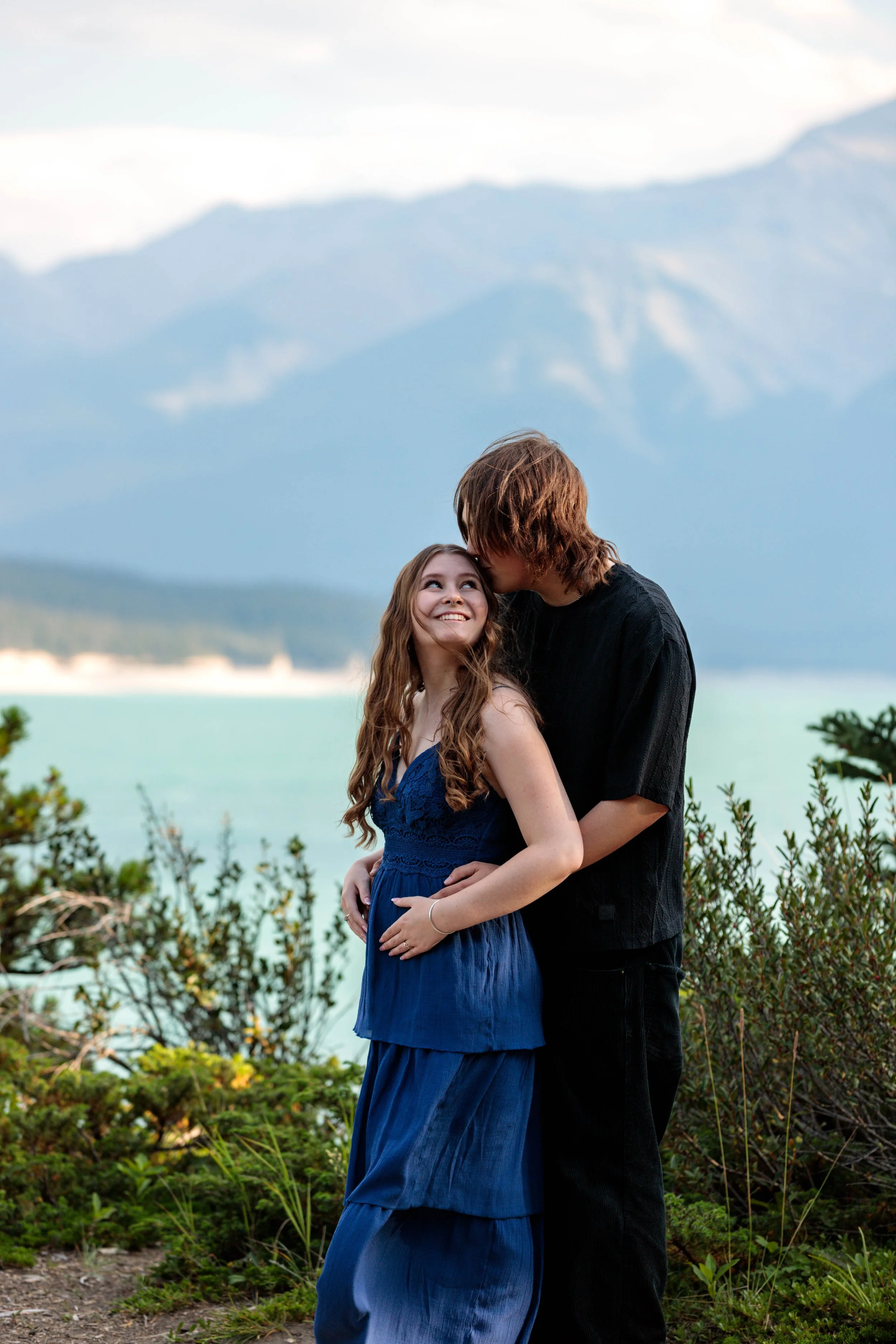 A young woman in a blue dress smiling at a man with long hair in black clothing, in a natural outdoor setting with mountains and a lake in the background.