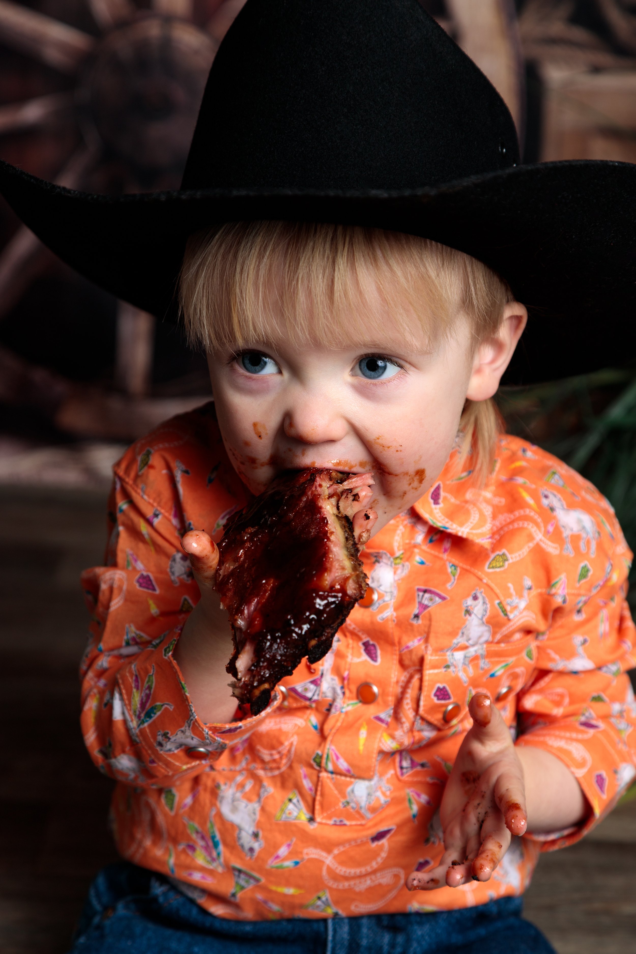 A young child with blue eyes and blonde hair wearing an orange shirt with a dog pattern, eating a large piece of barbecue ribs. The child has some sauce on their face and hands, and is sitting in front of a background with stacked logs.
