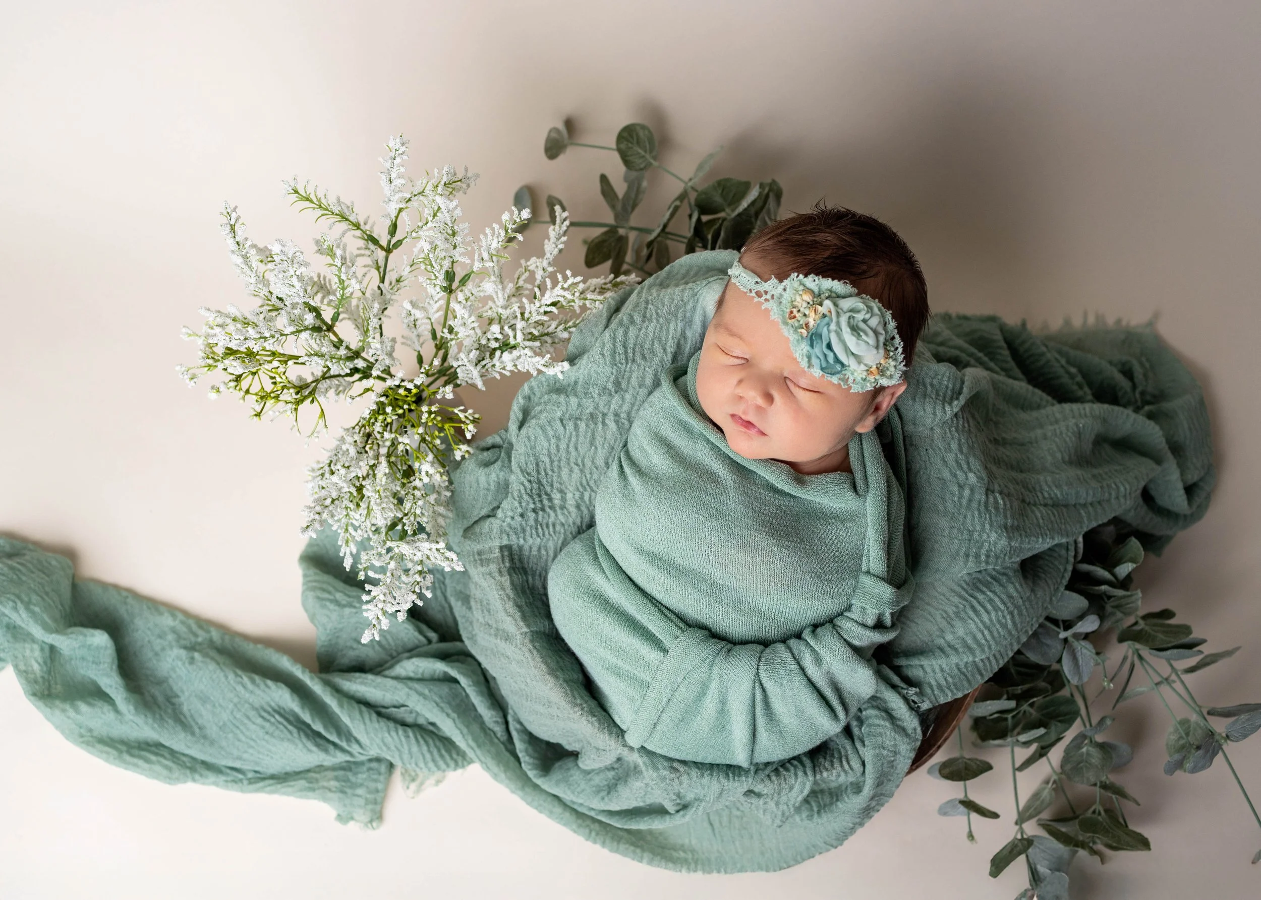 A peaceful baby with closed eyes wearing a floral headband, wrapped in a green blanket, lying on a cozy surface surrounded by green leaves and white flowers.