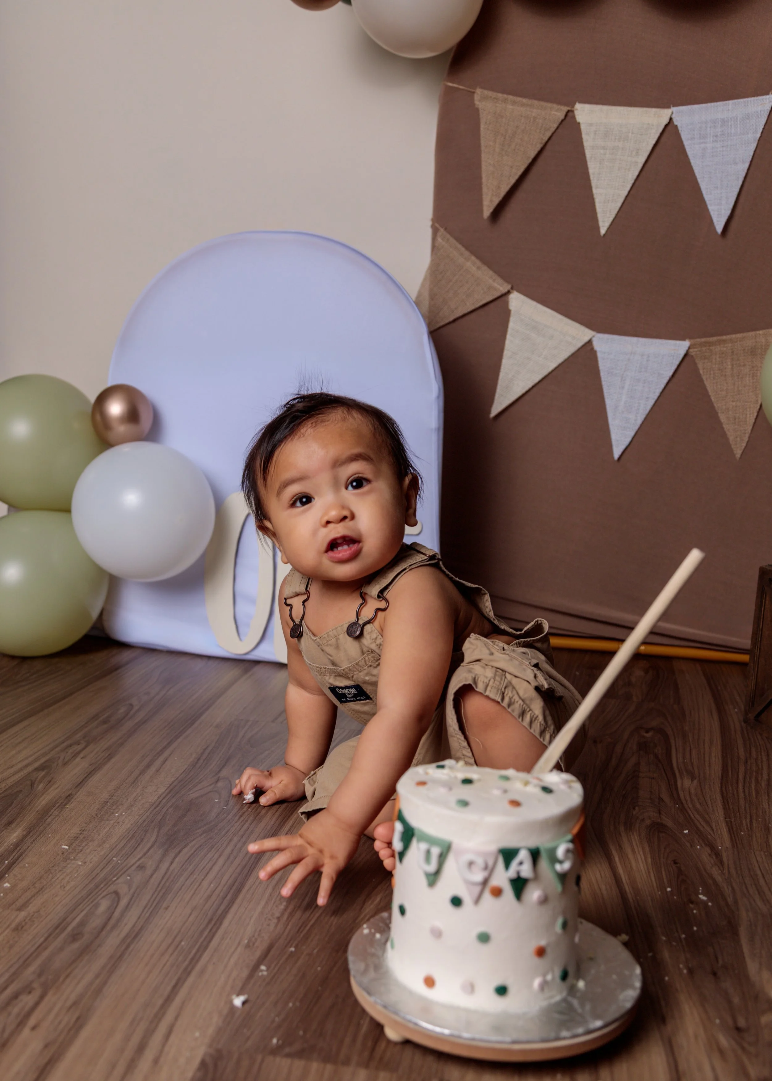 A young child with dark hair and wearing tan overalls is crawling on a wooden floor at a birthday celebration. In the foreground, there is a white cake with colorful sprinkles and a cake topper that seems to spell ' HAPPY BIRTHDAY' in small flags. Be