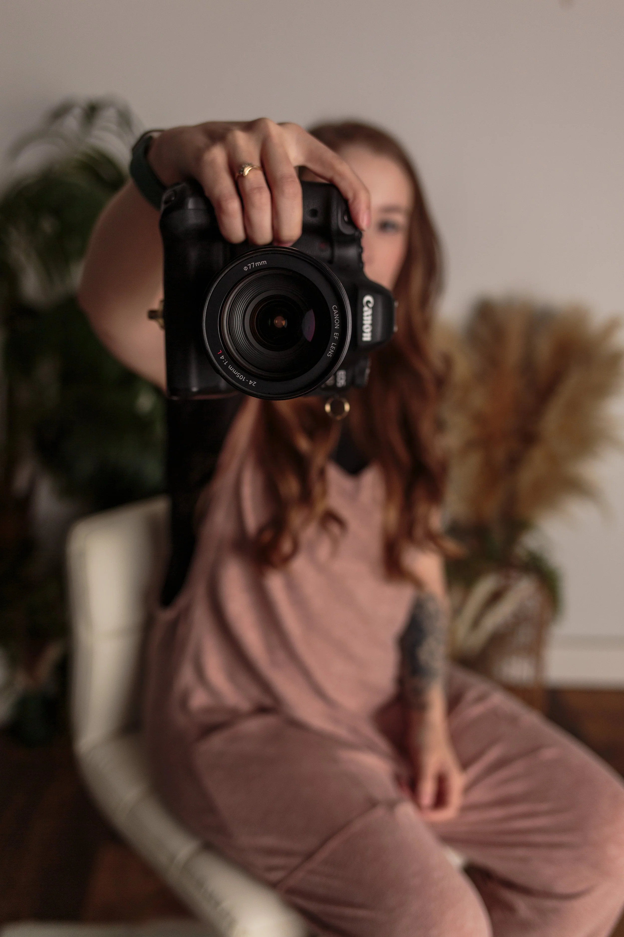 A woman with red hair taking a mirror selfie with a Canon camera. She is seated in a chair, wearing a pinkish-brown tracksuit, and has a ring on her finger. There are plants in the background.
