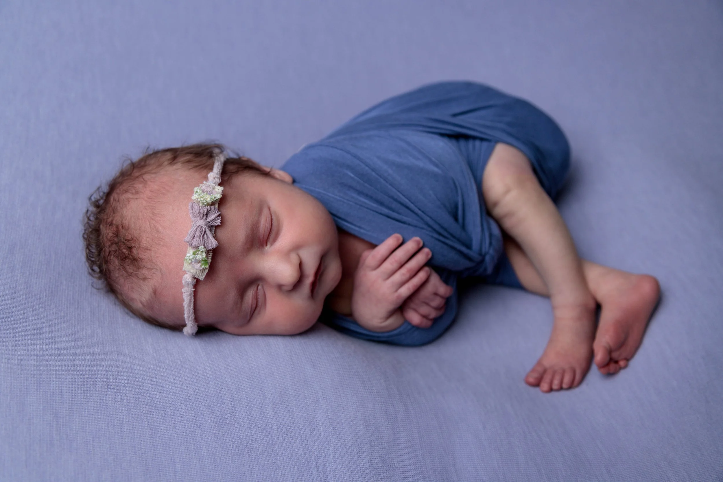 A sleeping newborn baby with a floral headband, lying on a light purple surface, with an adult's hand gently holding the baby's hand.