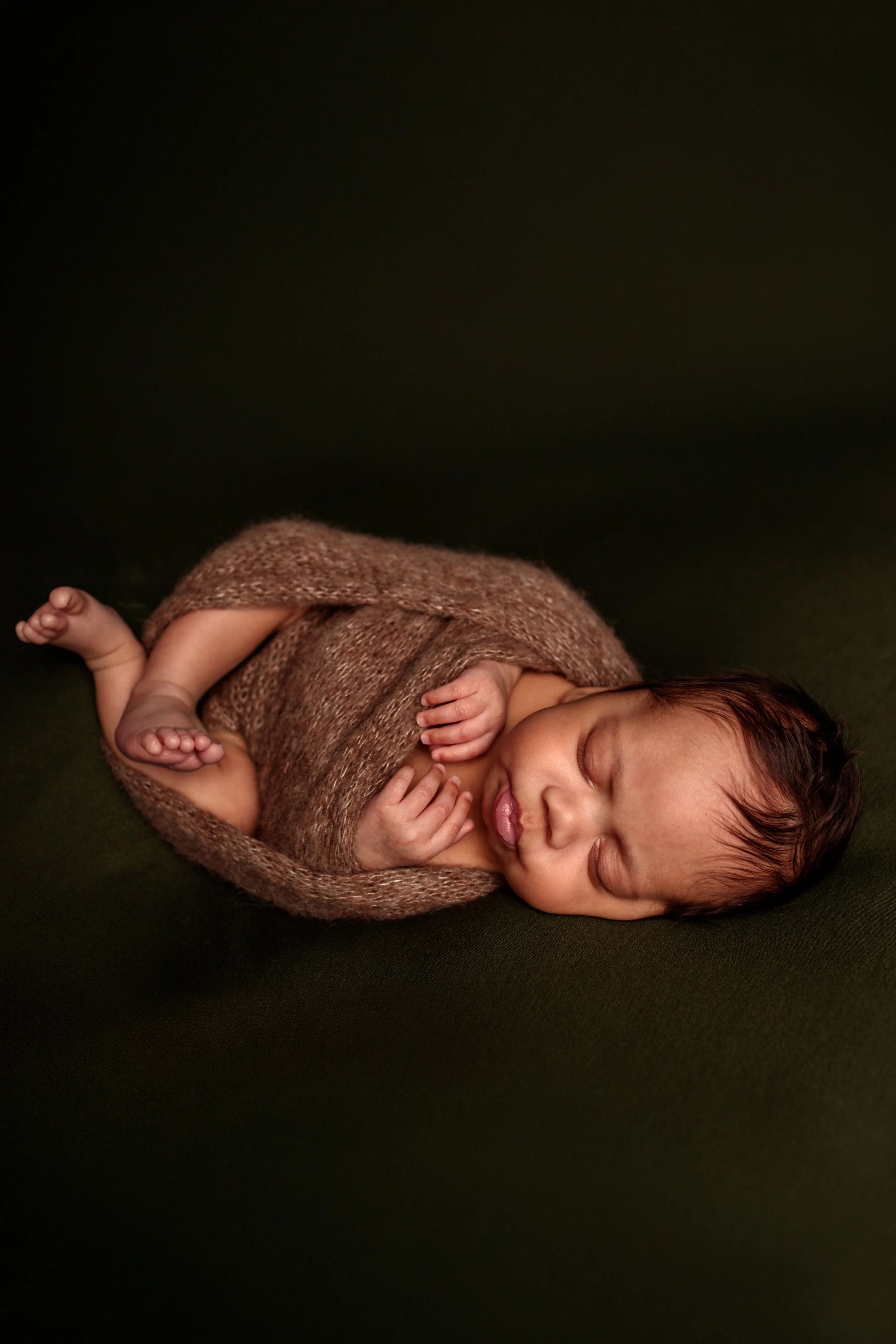 A newborn baby sleeping on a dark surface, wrapped in a brown knitted blanket, with a peaceful expression and tiny hands near the face.