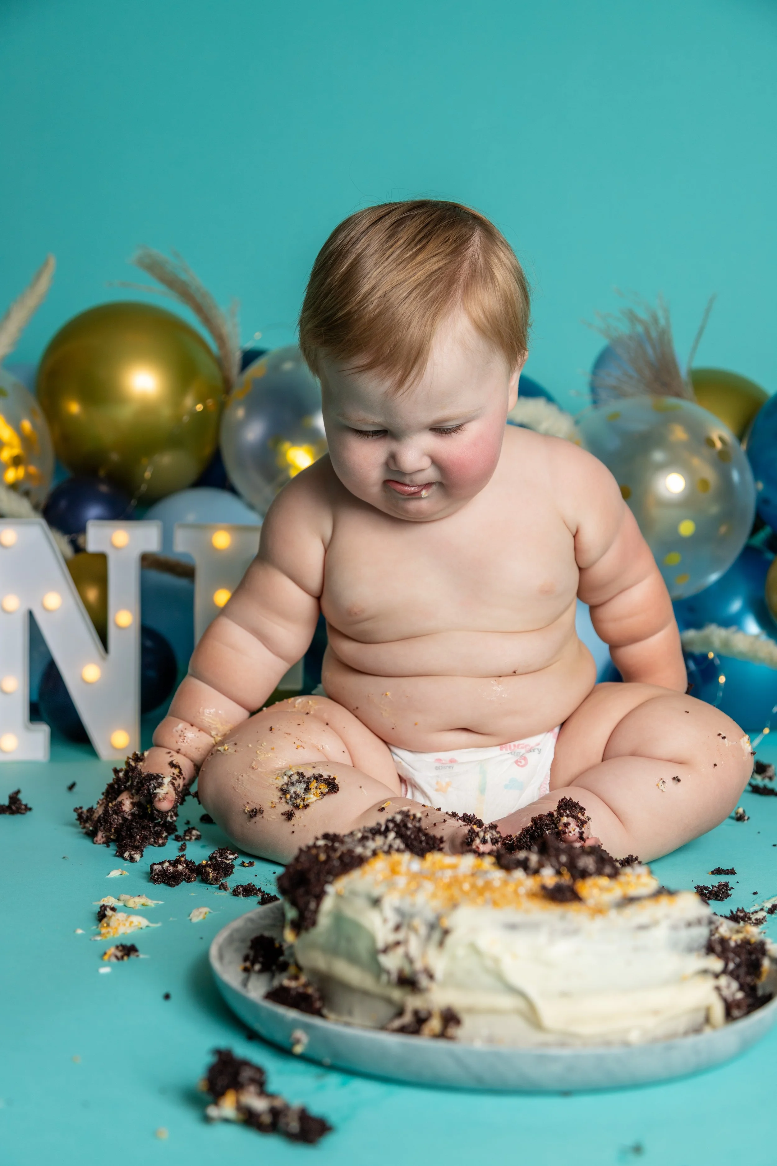 A young child with red hair and no shirt sits on the floor, surrounded by a mess of cake crumbs and icing, in front of a birthday cake with white frosting and darker crumbs. The background features gold and blue balloons and a teal wall, with decorative letters lit with small lights.