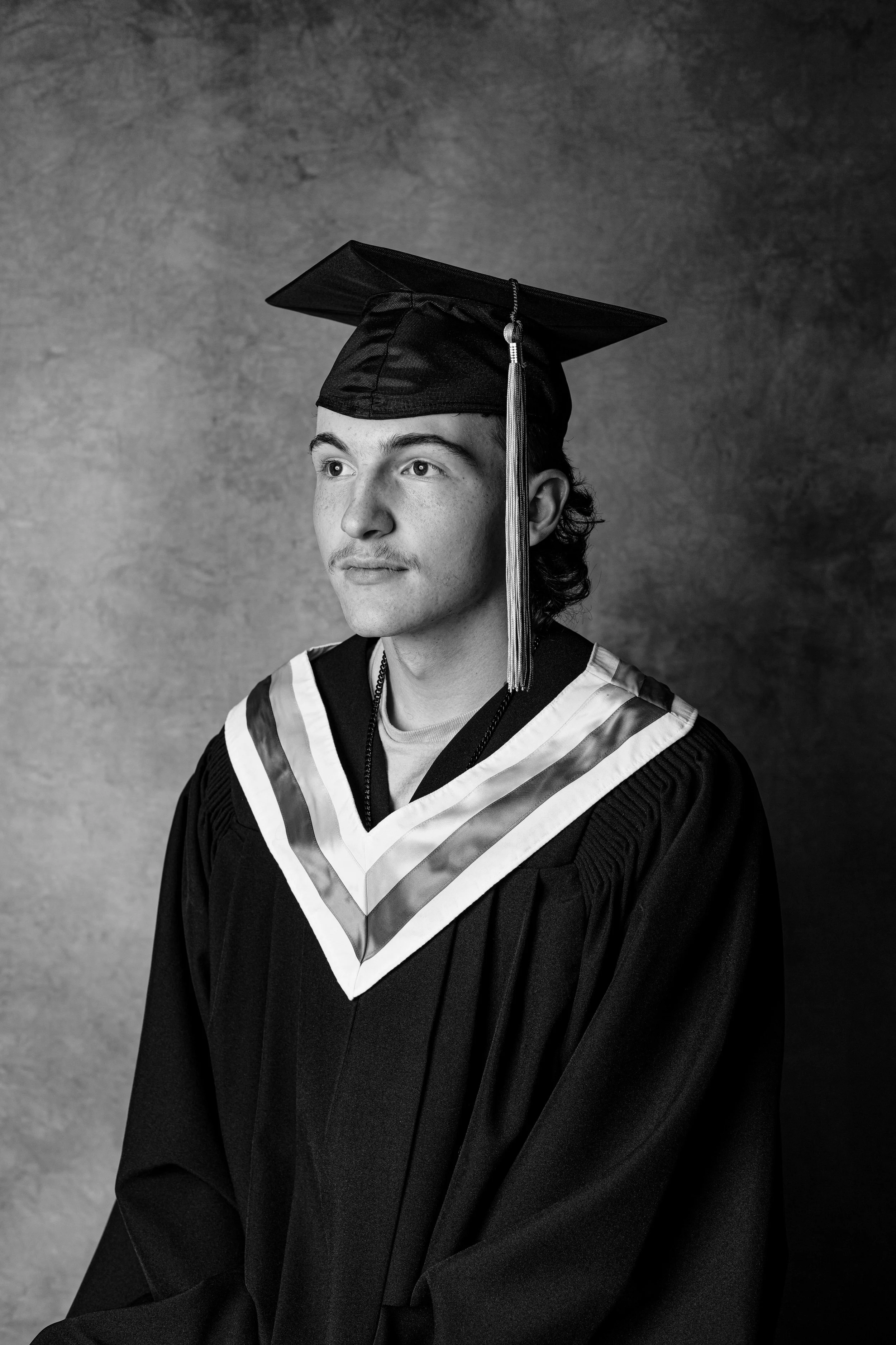 A young man in graduation cap and gown, looking to the side, in black and white photography.