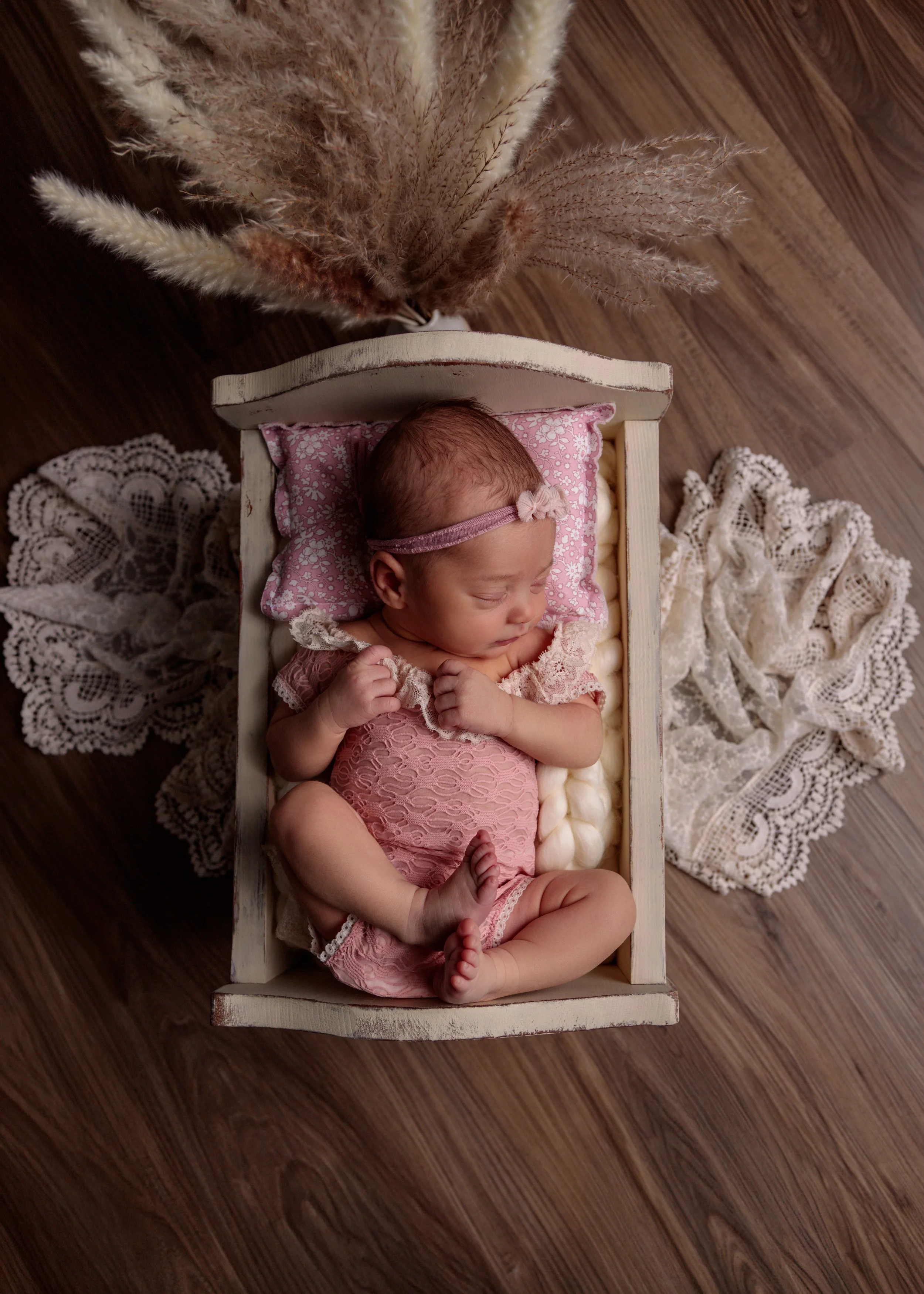 A newborn baby girl sleeping in a small white wooden bed, wearing a pink lace outfit and a pink headband with a bow, surrounded by lace and dried pampas grass on a wooden floor.