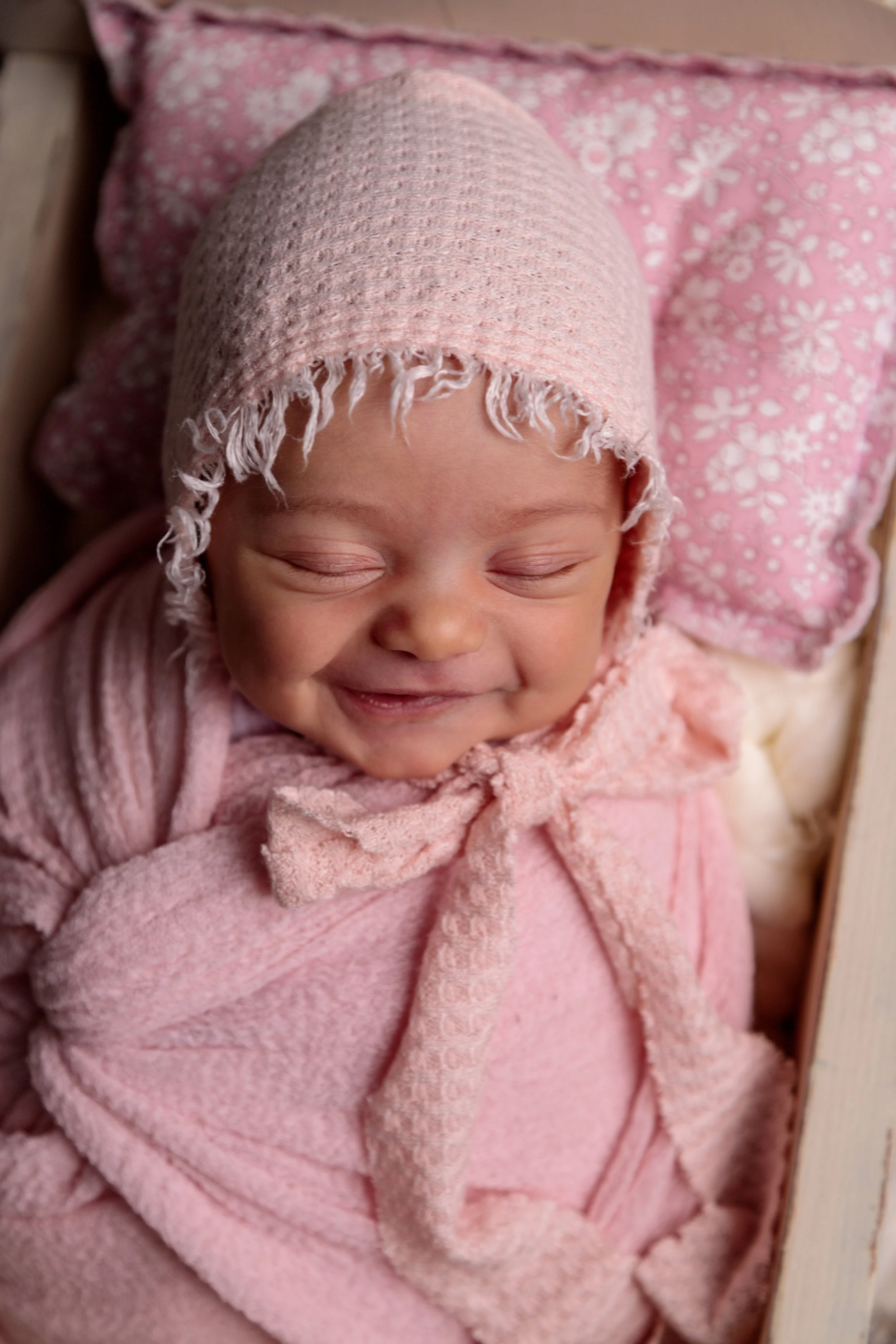 Close-up of a baby wearing a pink knitted hat and pink cozy clothing, smiling with eyes closed, lying on a pink floral pillow.
