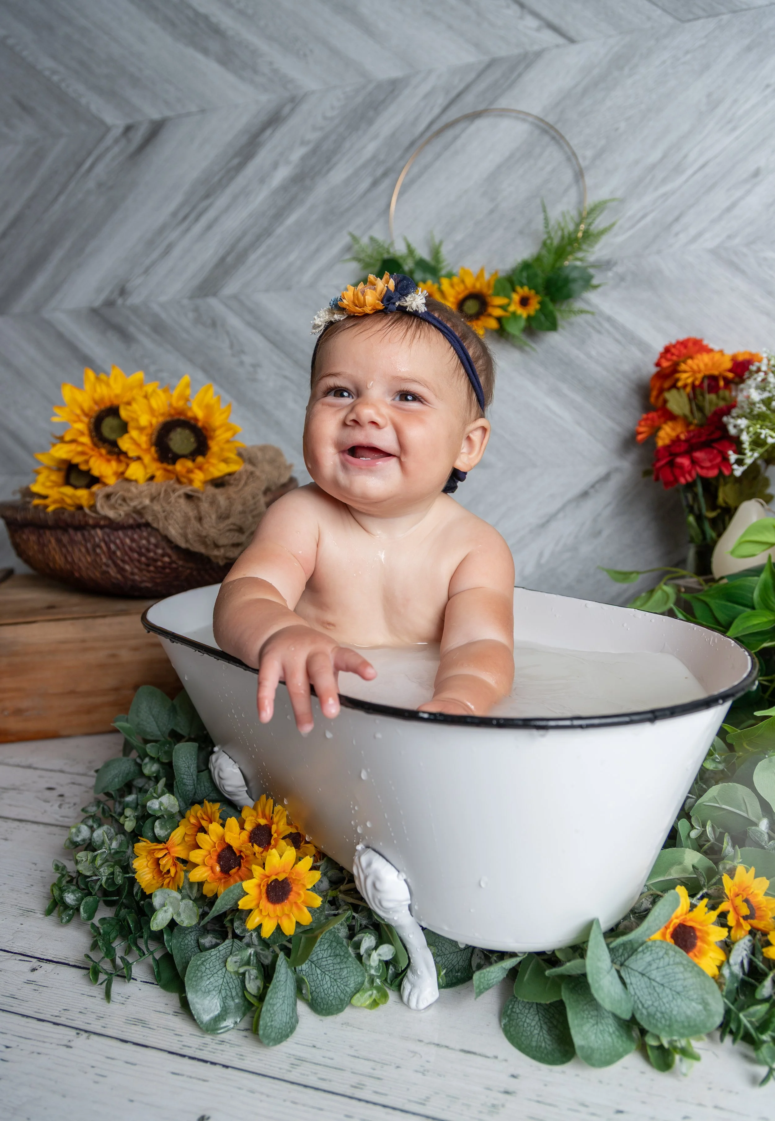 A smiling baby in a white bathtub surrounded by yellow sunflowers and green foliage, with a gray wooden wall background.