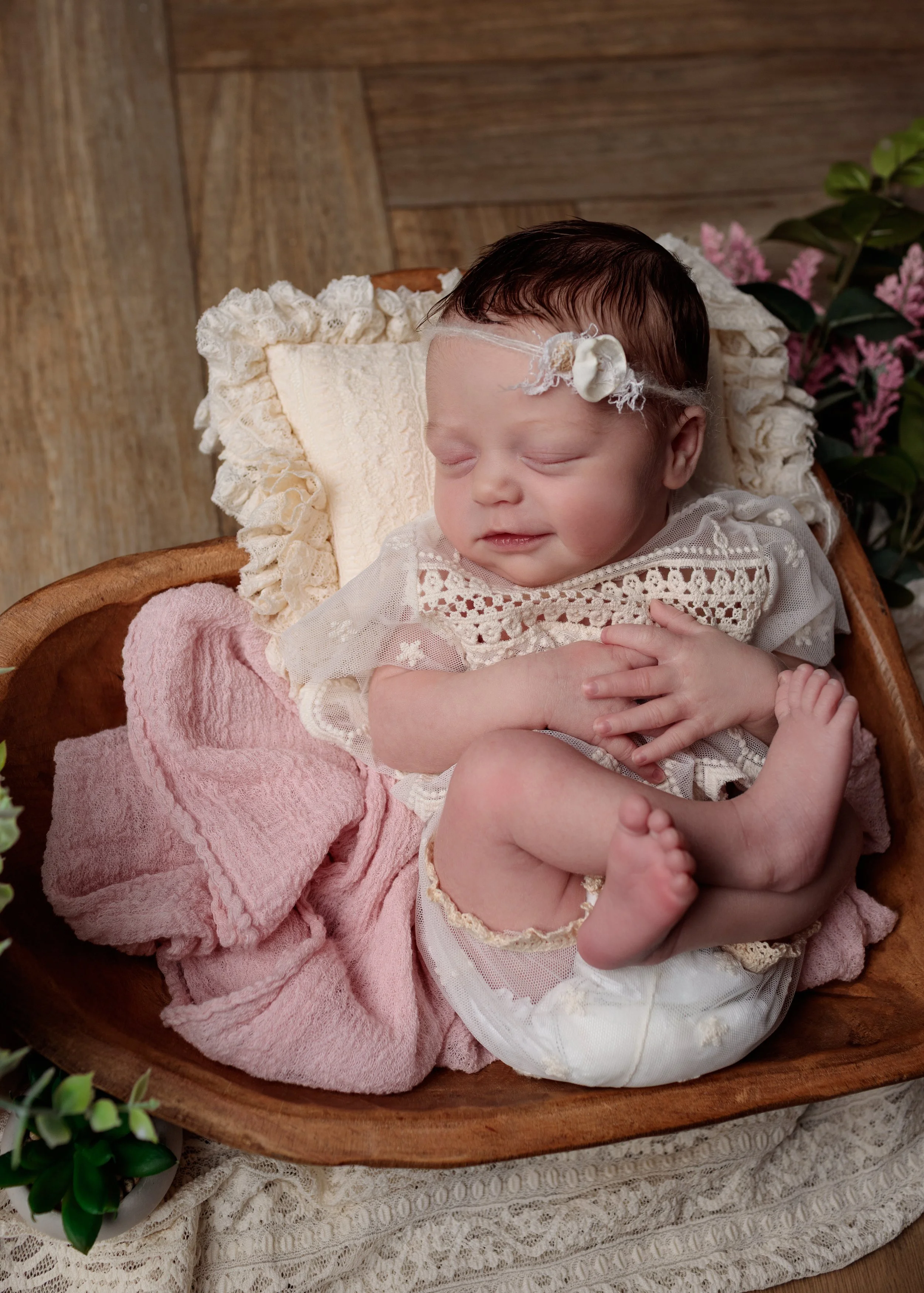 A newborn baby sleeping in a wooden cradle, wearing a white lacy outfit with a flower headband, surrounded by pink and green plants, on a lace-covered surface.