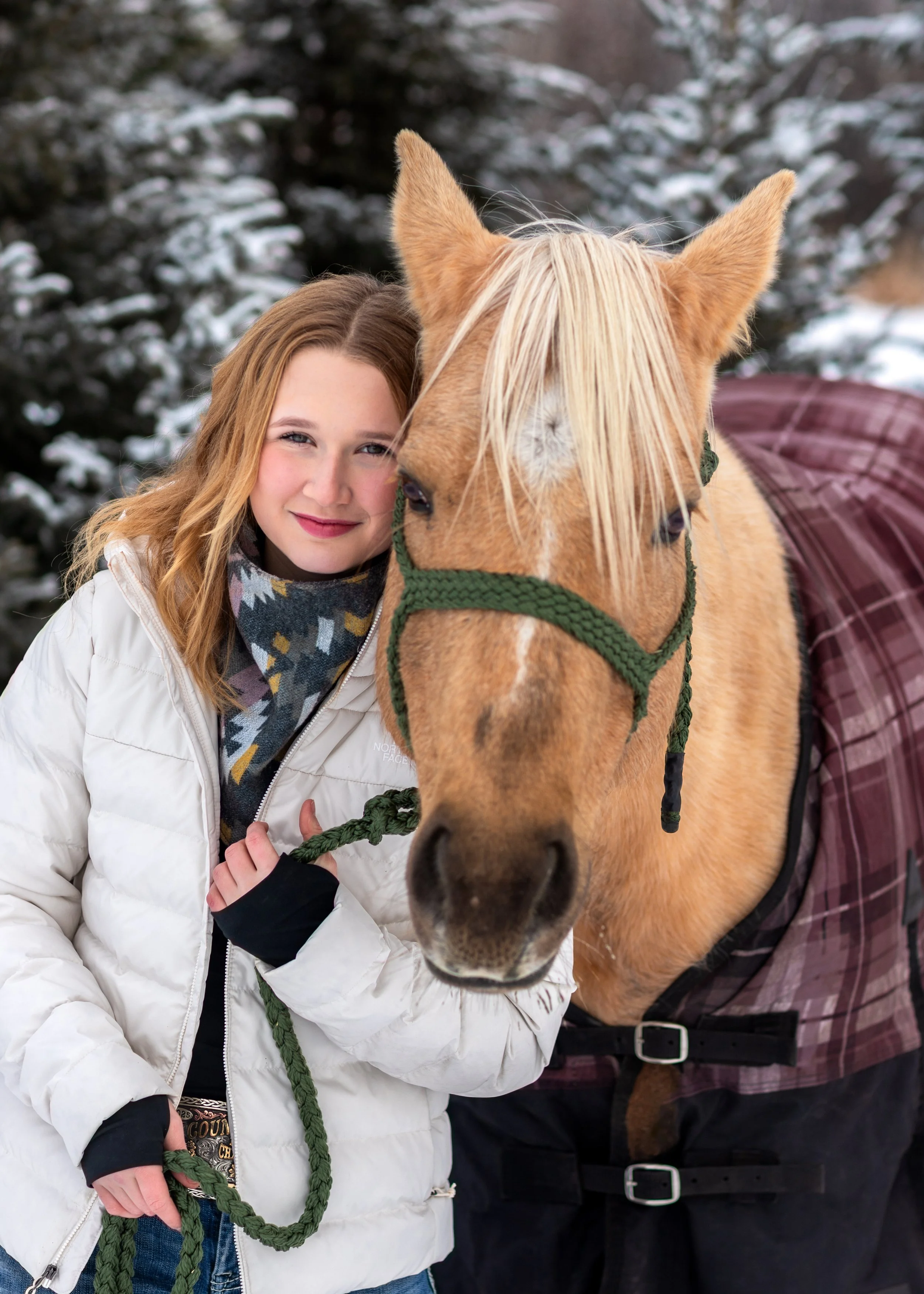 A woman with blonde hair and a white jacket holding a green lead, standing next to a brown horse with a blonde mane, in a snowy outdoor setting.