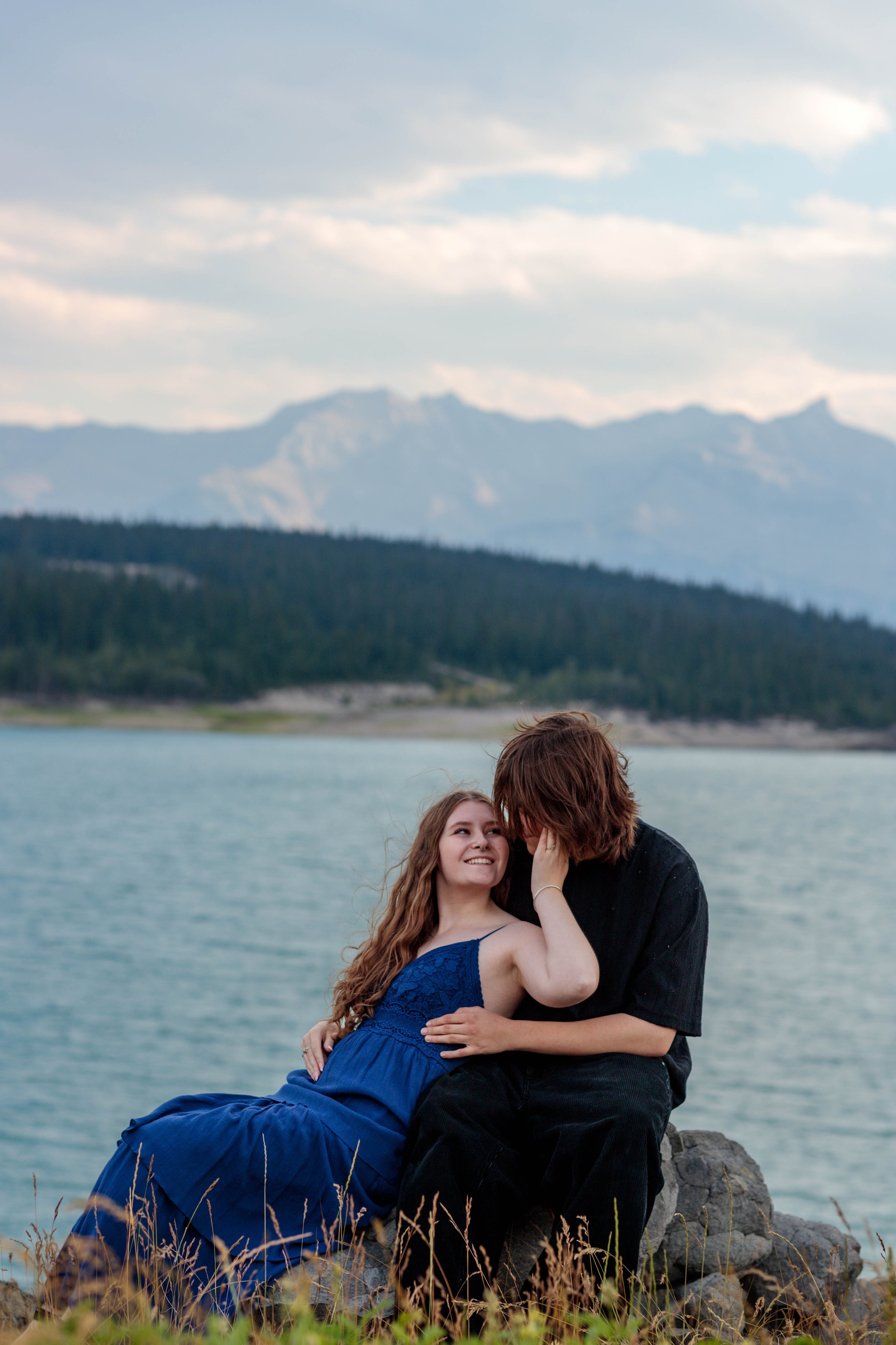 A young woman in a blue dress and a young man in dark clothing sitting on rocks by a body of water, with mountains and trees in the background, sharing an intimate moment.