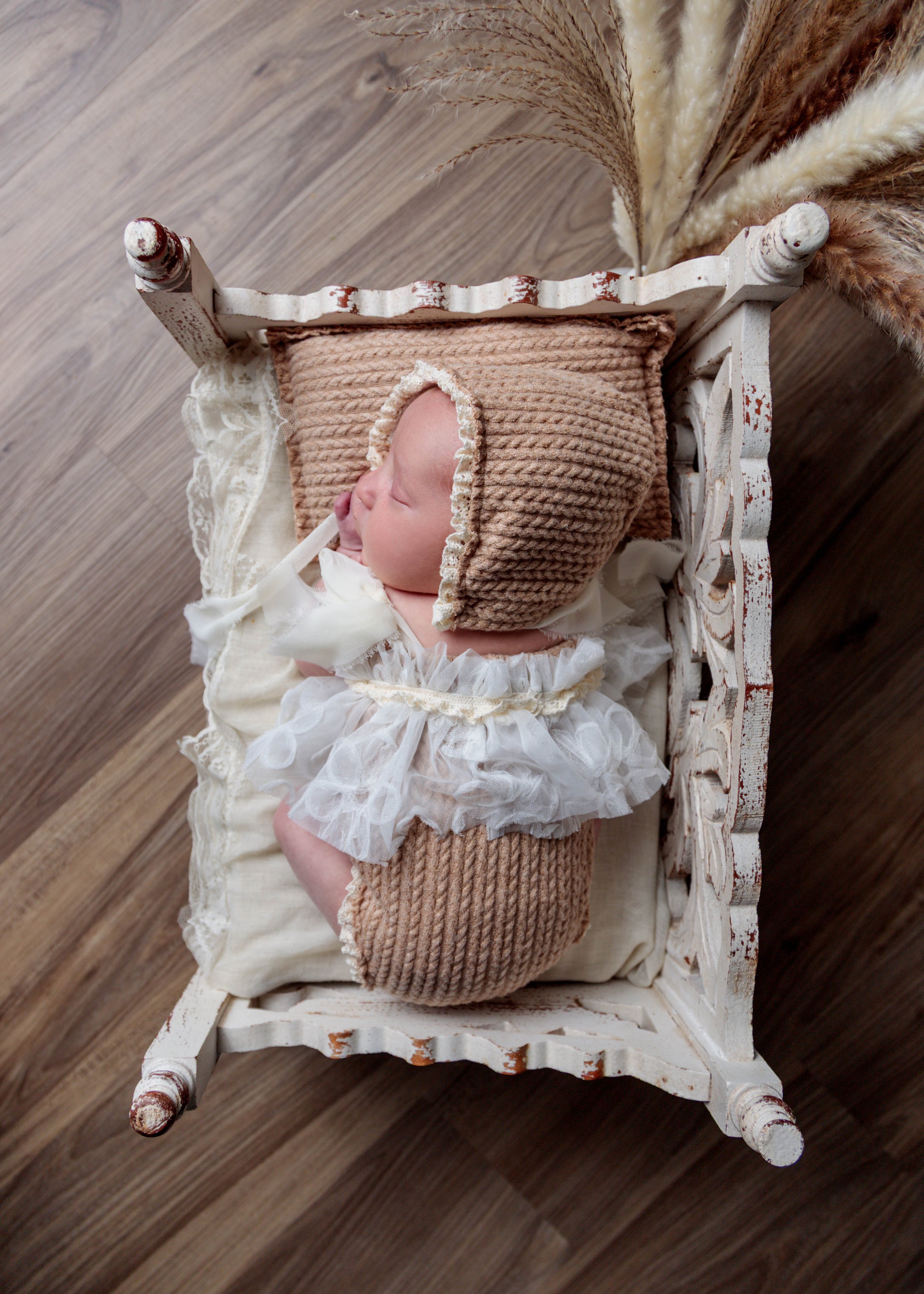 A sleeping baby in a vintage white wooden cradle with distressed paint. The baby is wrapped in a beige corduroy blanket and wearing a frilly, cream-colored dress and bonnet. Dried pampas grass is placed behind the cradle.