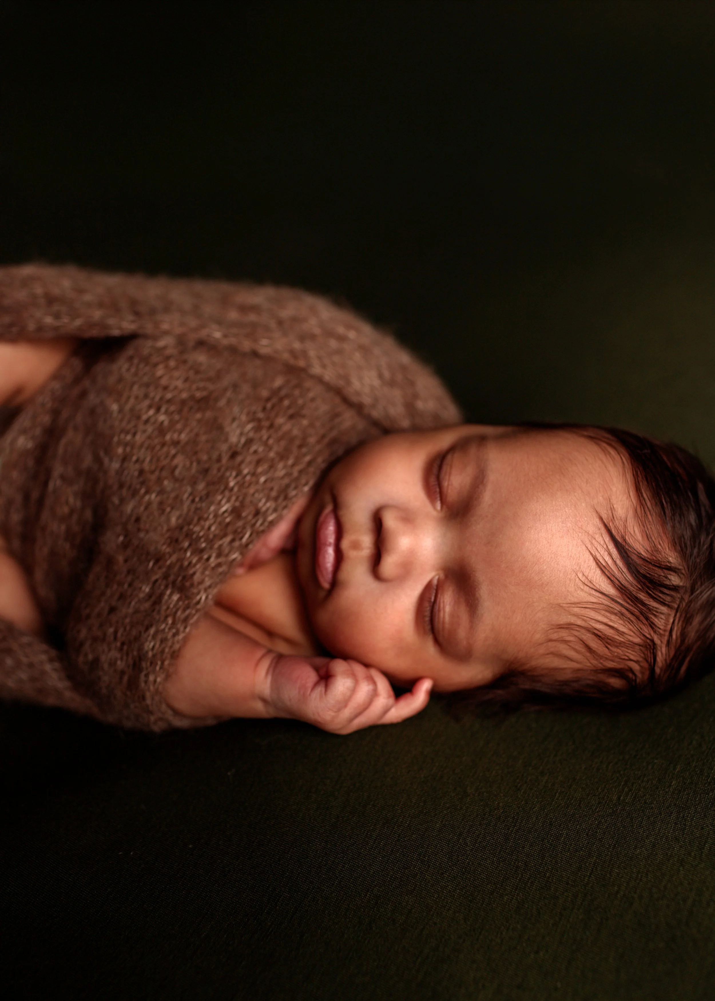 A close-up of a sleeping baby with dark hair, lying on a black surface, wearing a brown textured sweater, with a peaceful expression.