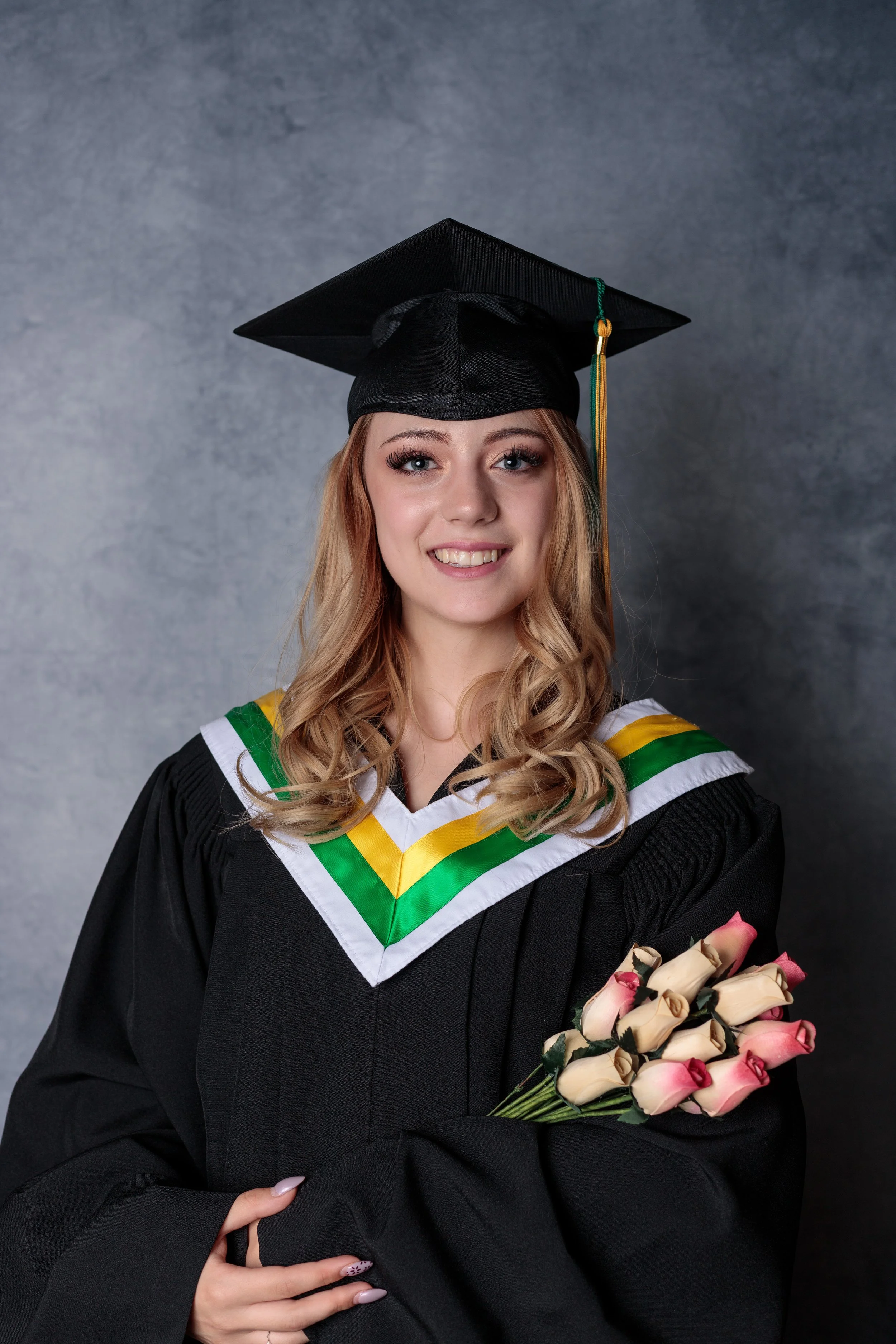 A young woman in a graduation cap and gown holding a bouquet of pink and cream roses.