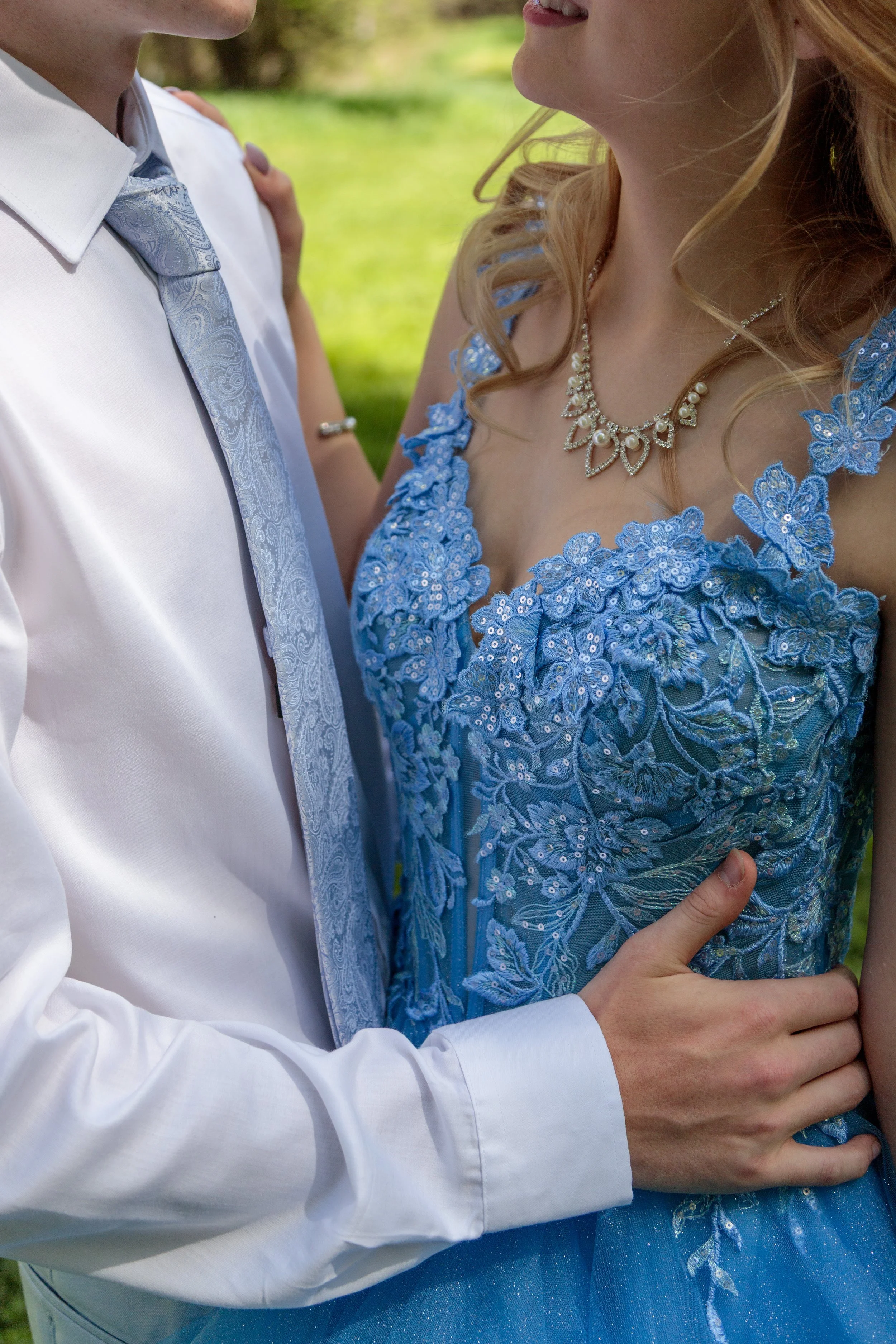 Close-up of a couple wearing formal attire embracing outdoors. The woman wears a blue embroidered dress and a pearl necklace; the man is in a white shirt with a patterned tie. Their faces are partially visible, capturing an intimate moment.