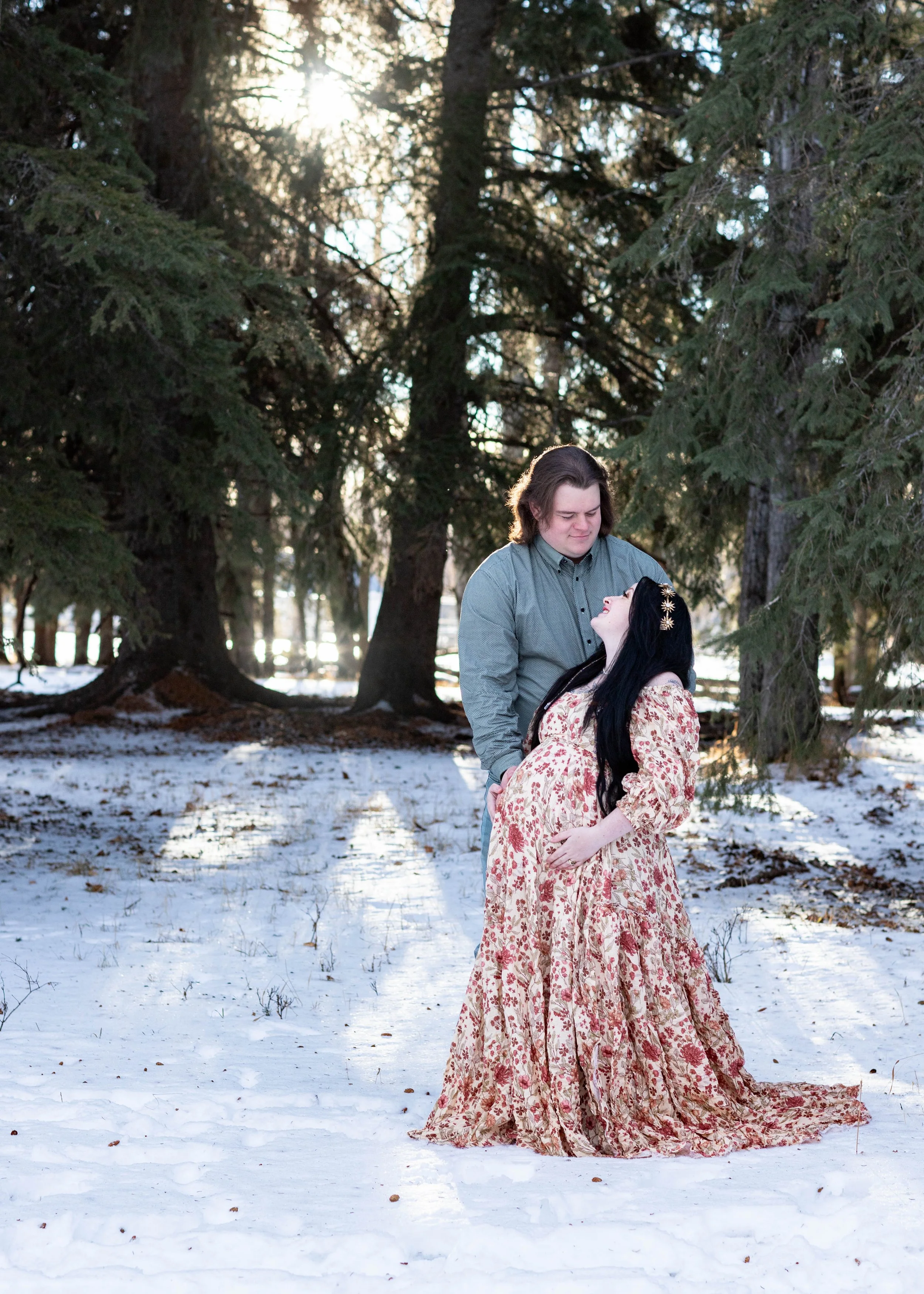 A man and a woman standing in a snow-covered forest with tall evergreen trees. The man, with shoulder-length brown hair, is wearing a gray shirt and is looking down at the woman. The woman, with long black hair adorned with gold leaf clips, is wearing a flowing floral dress and is looking up at the man. The sunlight filters through the trees in the background.