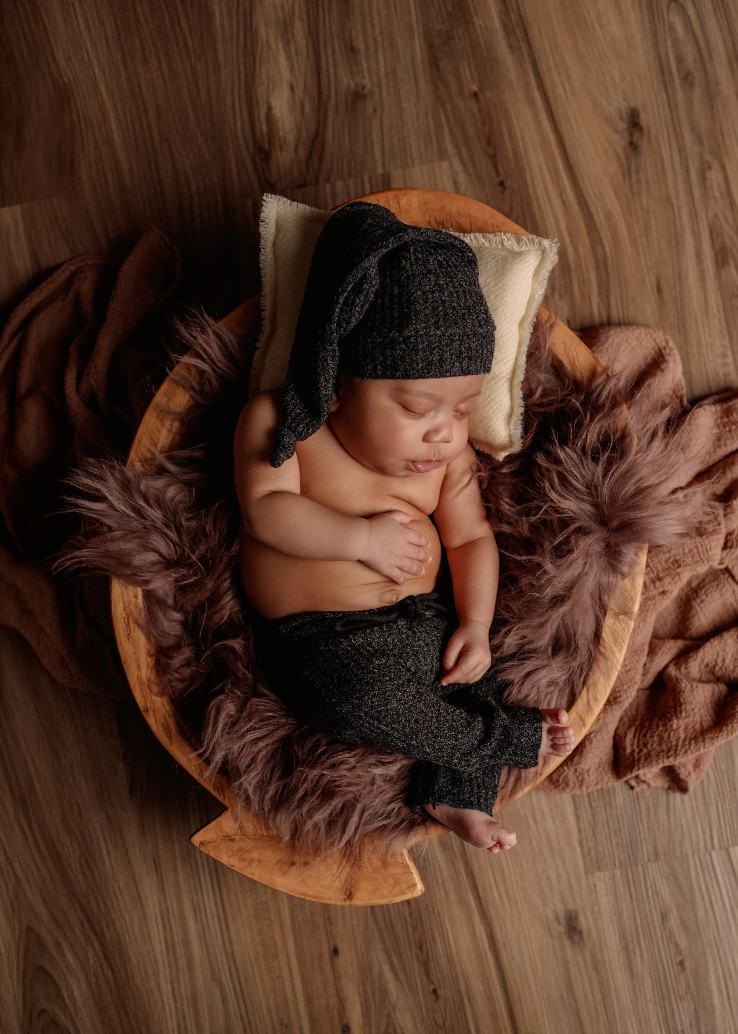 A baby sleeping on a small round wooden bed with plush blanket, wearing a black knit hat, surrounded by soft blanket and brown fur bedding.