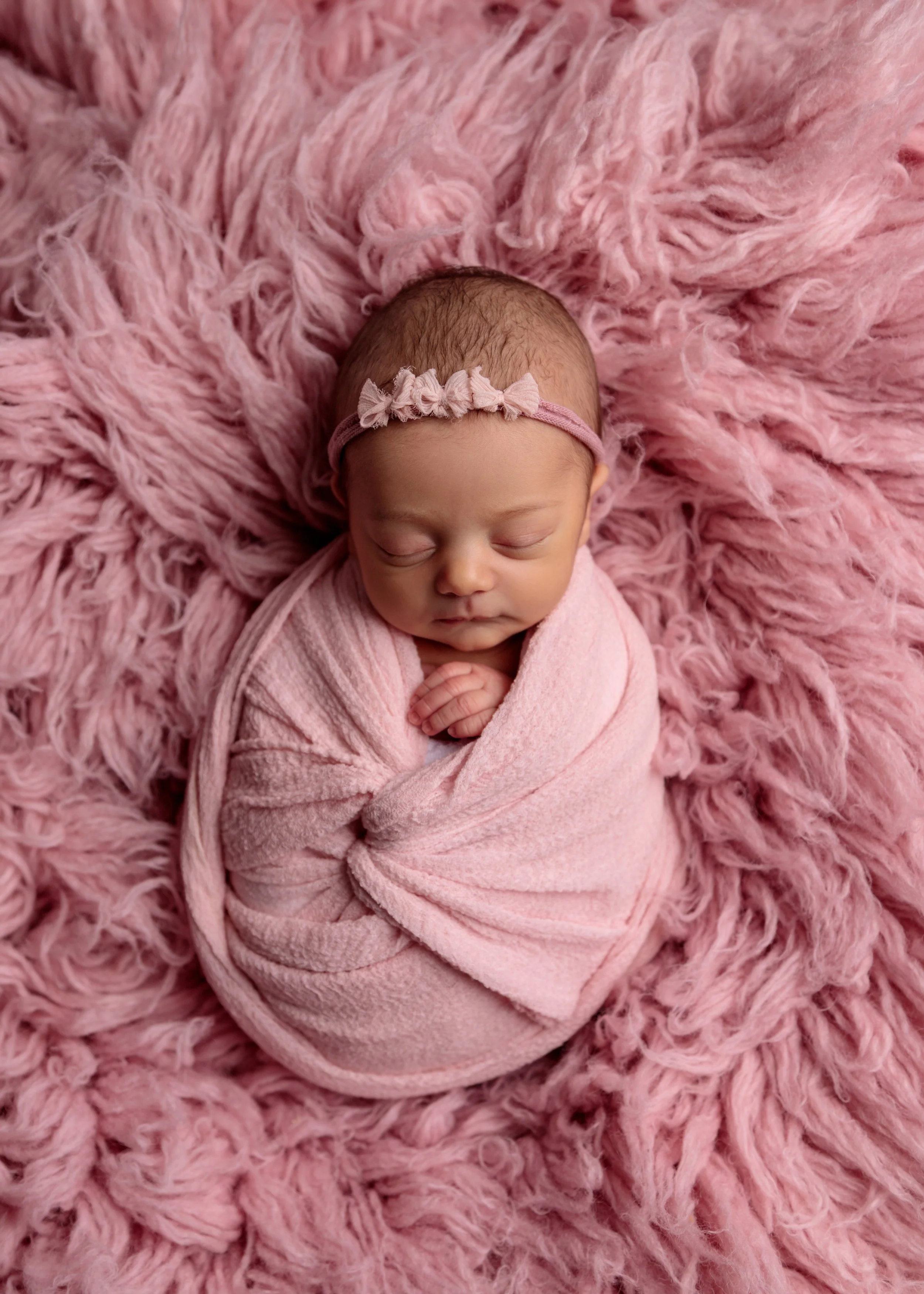 A newborn baby peacefully sleeping on a fluffy pink blanket, wrapped in a soft pink blanket, with a matching pink headband with a bow.