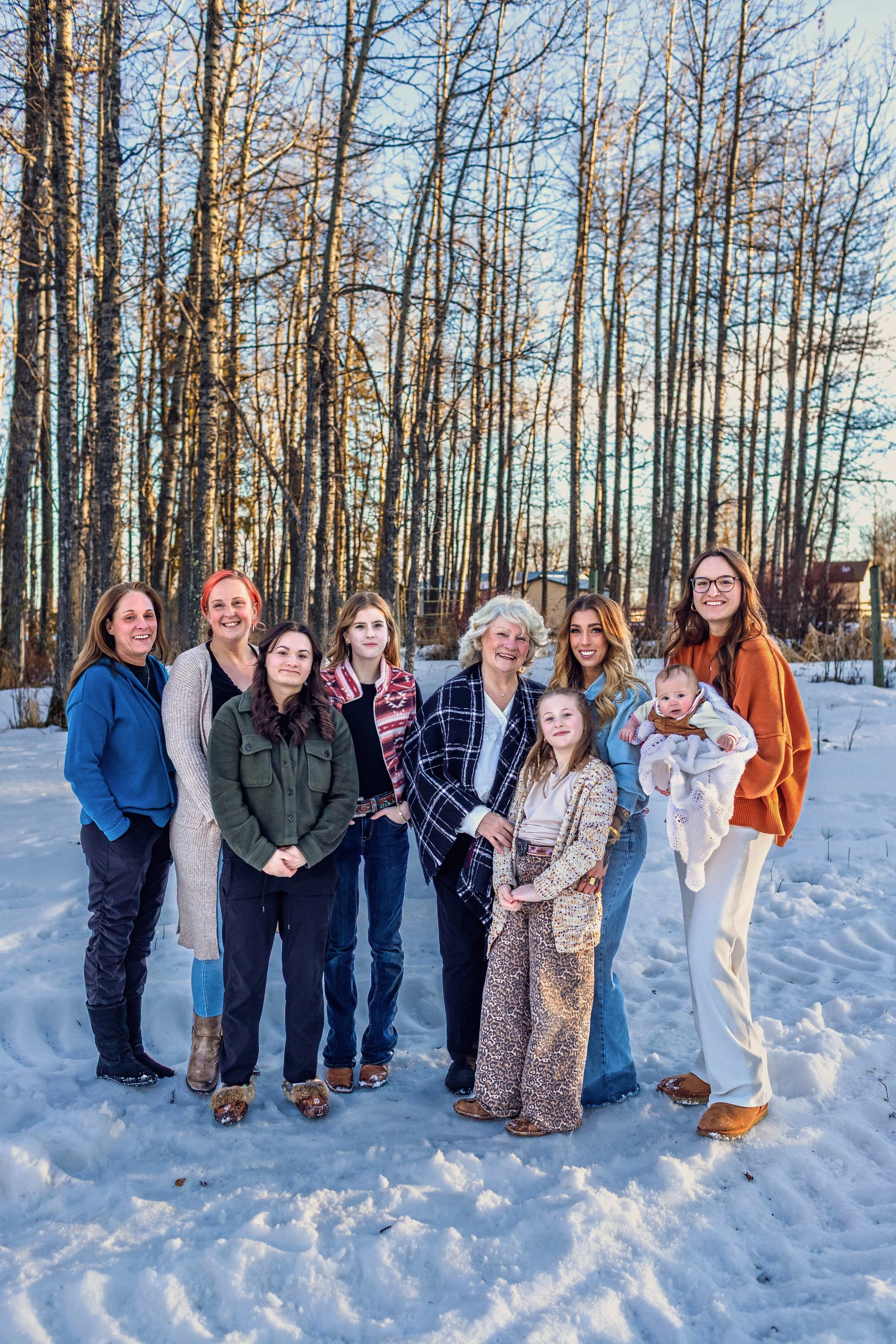 Group of women and children standing outdoors on snow-covered ground with bare trees and houses in the background, smiling at the camera.