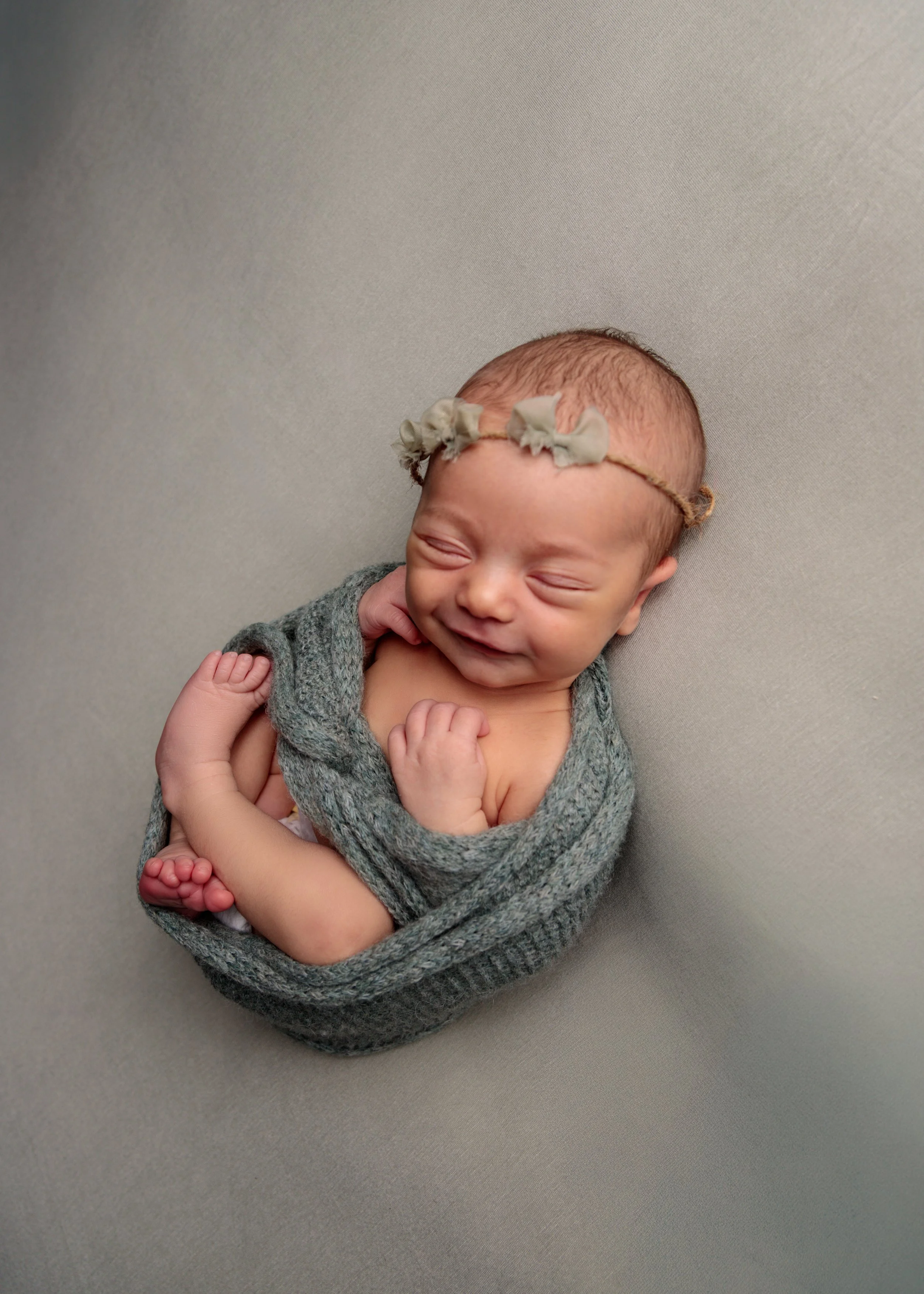 A newborn baby girl with a floral headband, dressed in a gray knitted outfit, lying on a soft surface with a happy expression.