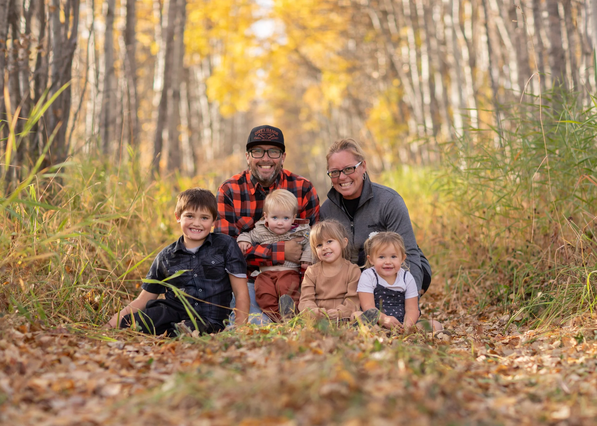 Family portrait of seven people, including two adults and five children, sitting outdoors in a forest during autumn, surrounded by fallen leaves and trees with yellow leaves.