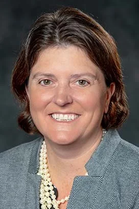 A woman with short brown hair, wearing a gray blazer and pearl necklace, smiling at the camera.