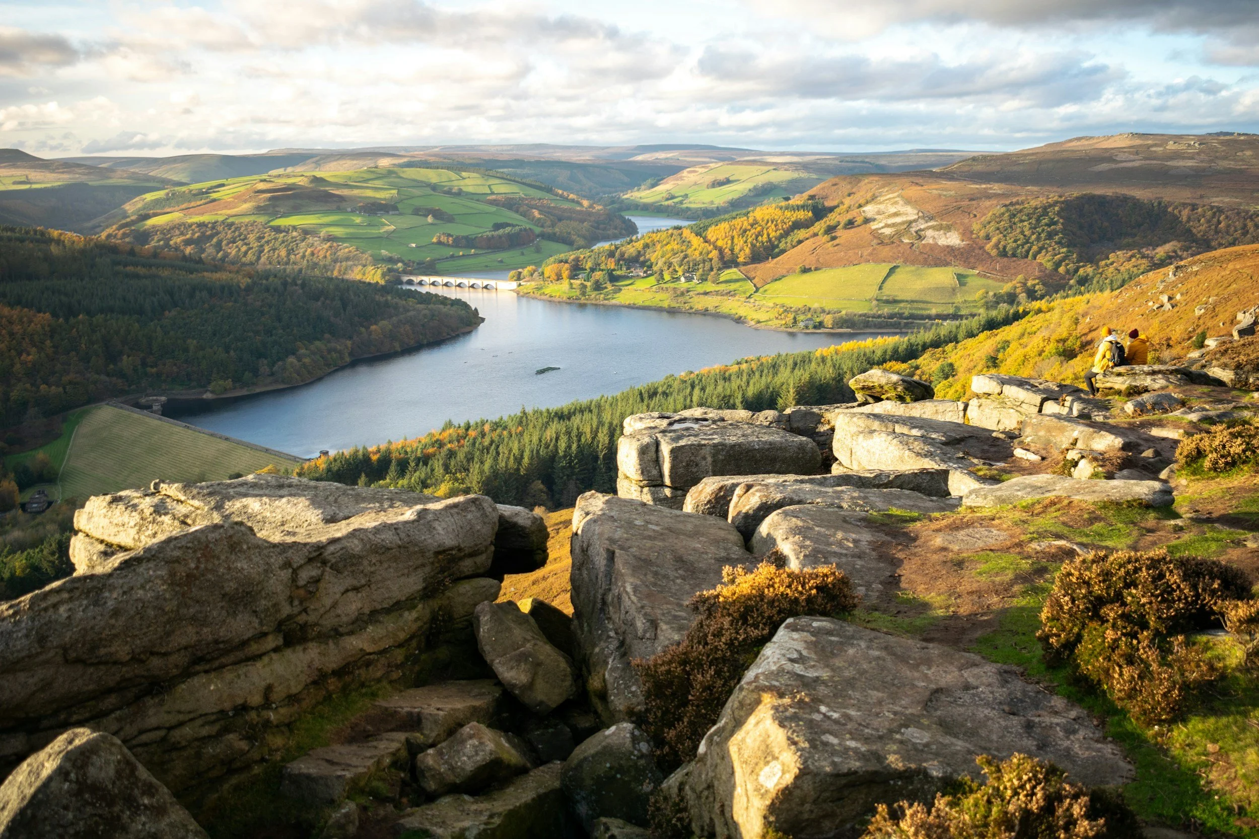 Scenic landscape of Ladybower Reservoir with stone foreground, surrounded by green hills, forests, and a distant bridge under a partly cloudy sky.