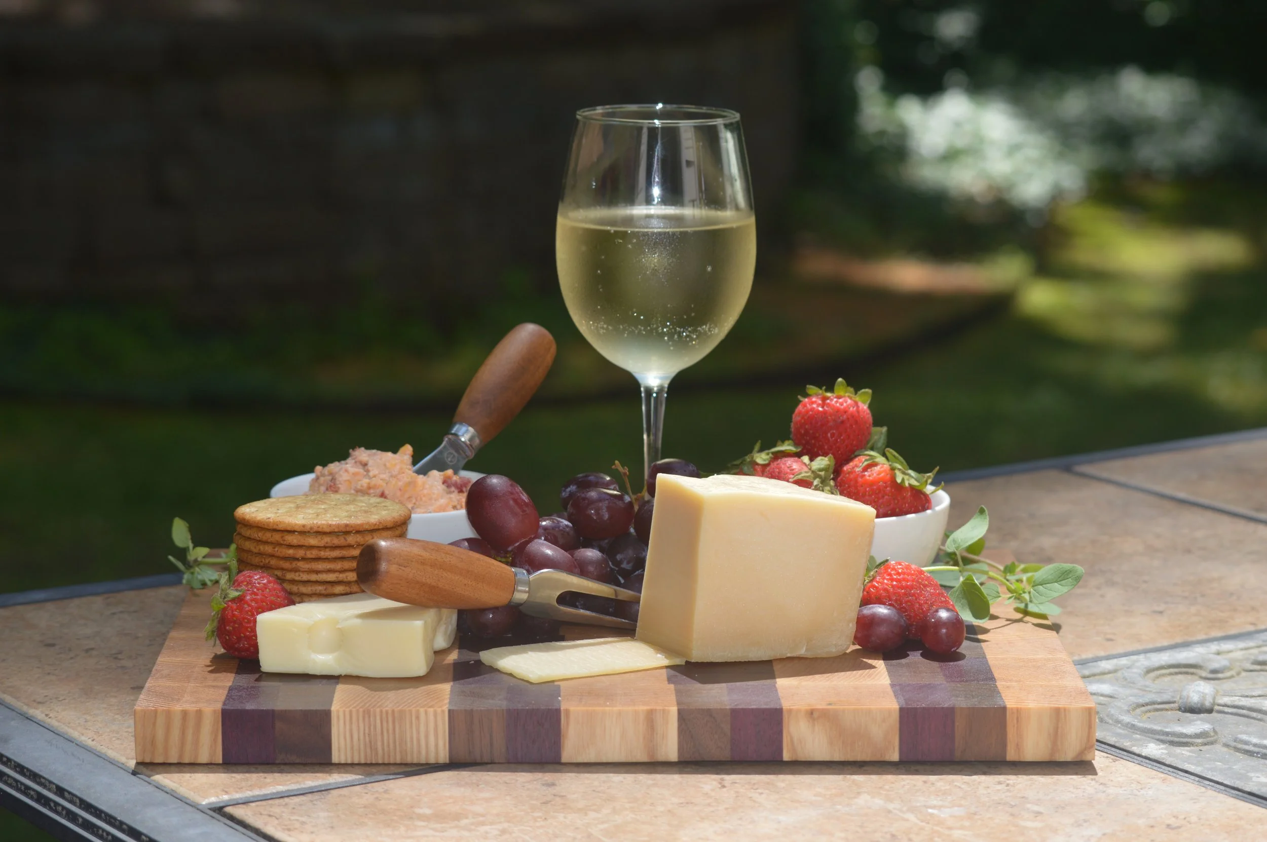 A cheese platter with grapes, strawberries, crackers, cheese, and a glass of white wine on a wooden cutting board outdoors.