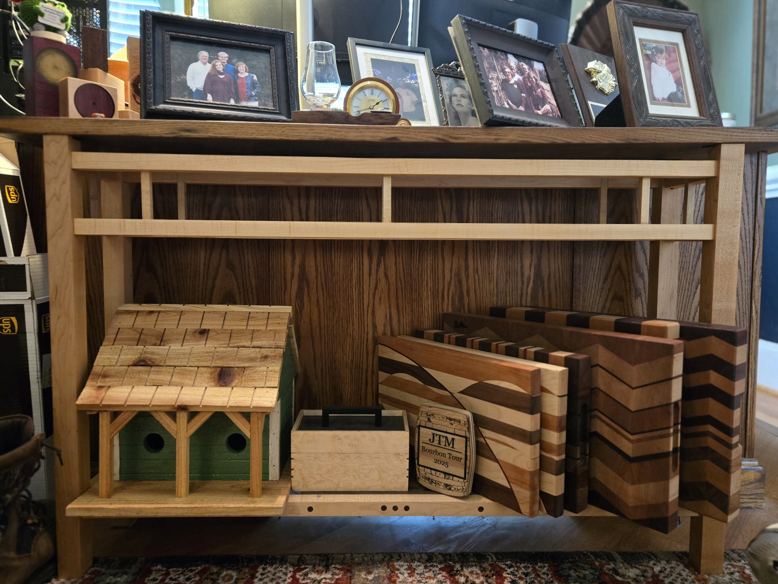 Display of handcrafted wooden items, including a miniature birdhouse, cutting boards with striped and geometric patterns, and other wooden accessories, stored on and behind a wooden table with framed photos and decorative objects on top.