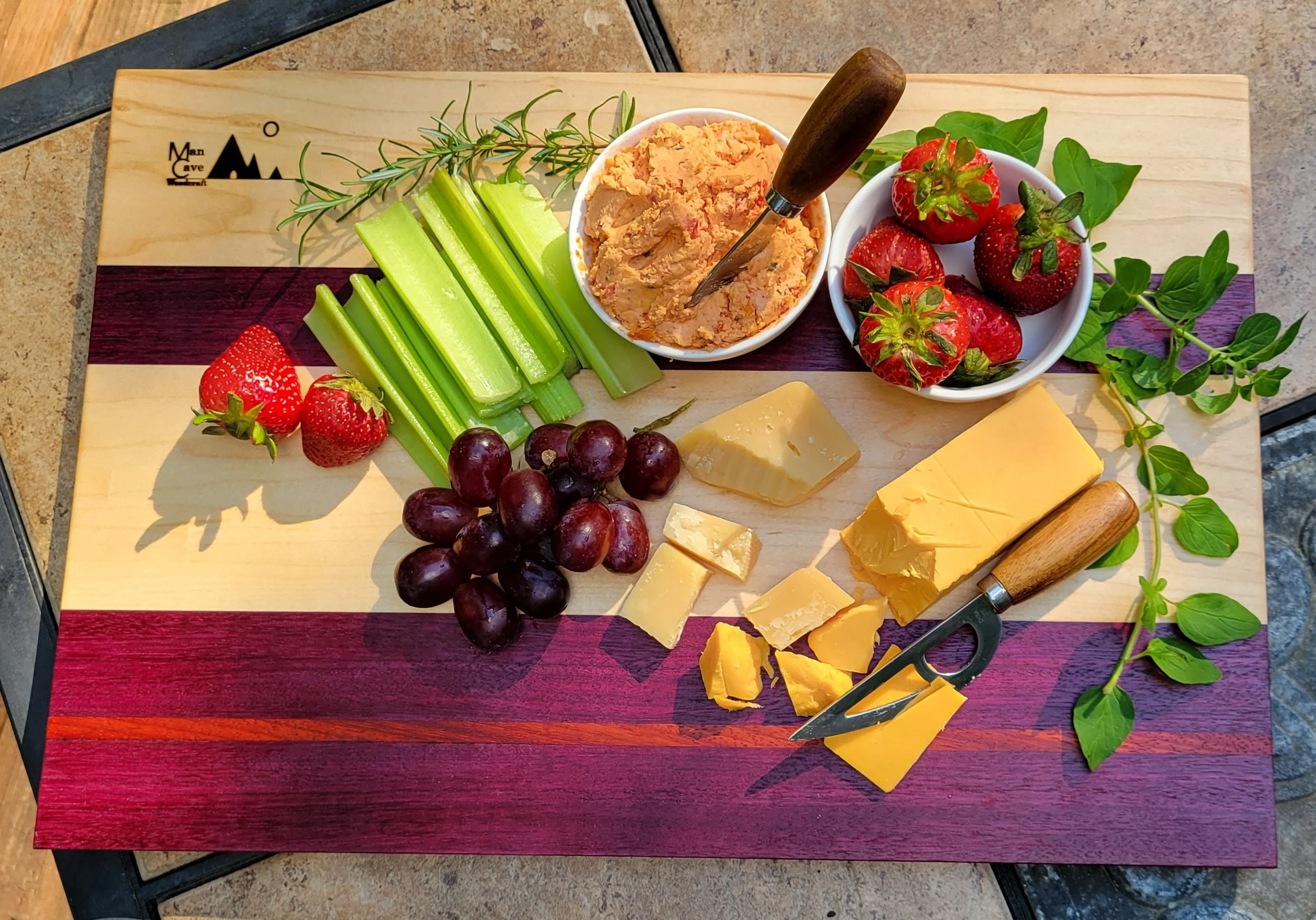 A charcuterie board with strawberries, grapes, celery sticks, cheese, hummus, and herbs on a wooden surface.