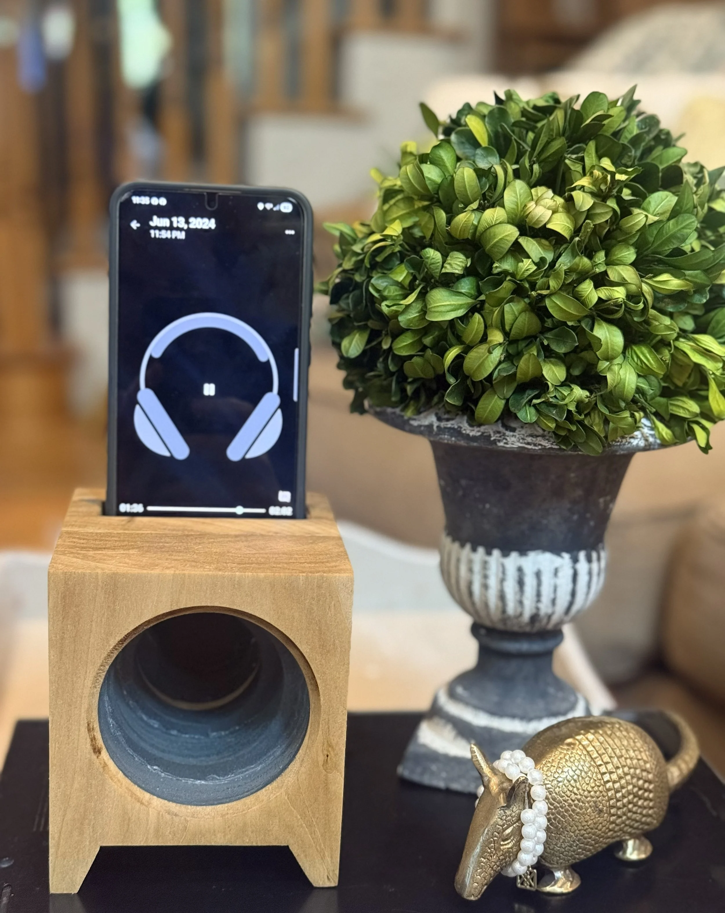 A wooden speaker holding a smartphone playing music with a headphone icon, a decorative potted green plant, a gold elephant figurine with a pearl necklace, and a black and white decorative vase in the background.