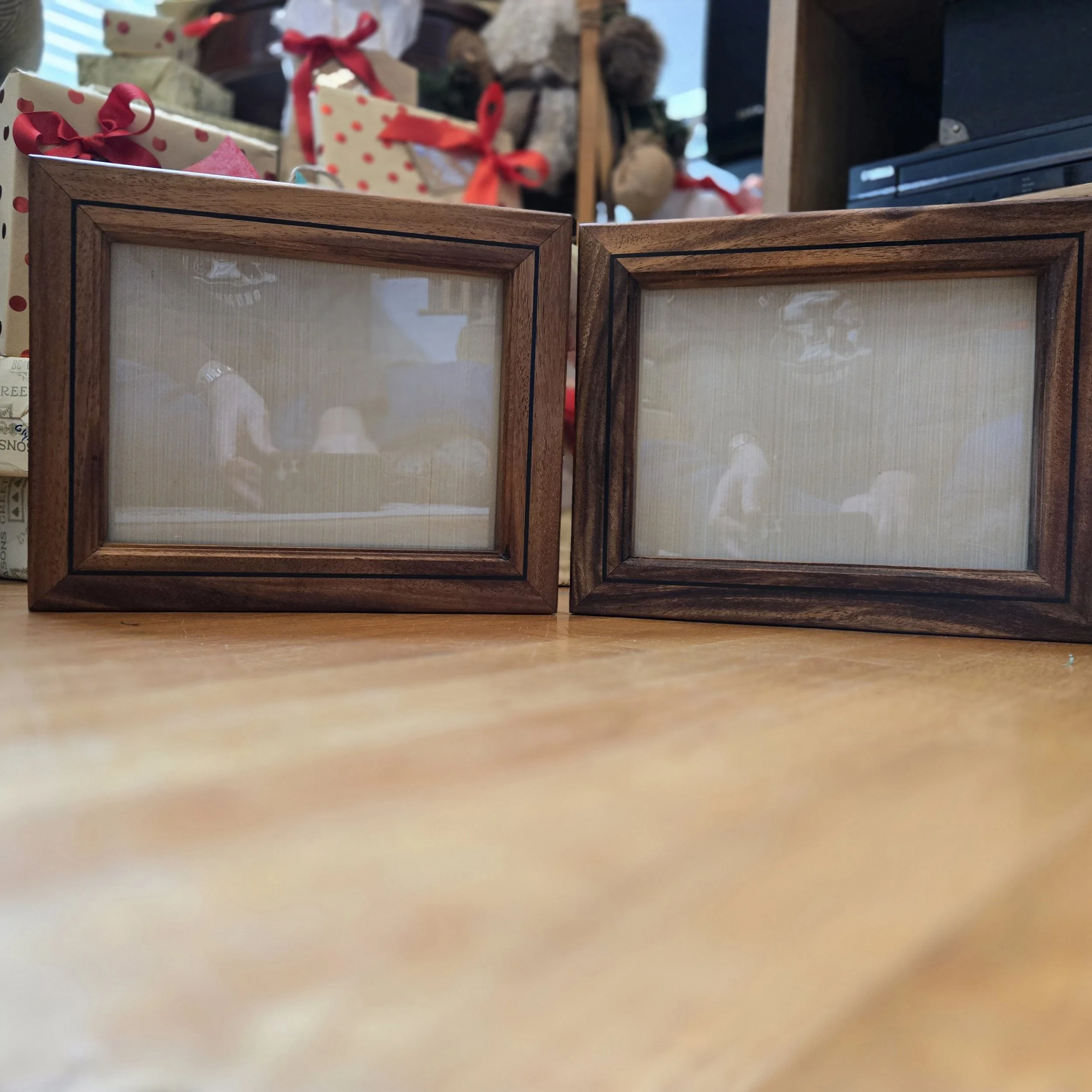 Two empty wooden picture frames with glass fronts on a wooden surface, with Christmas gift bags and stuffed animals in the background.