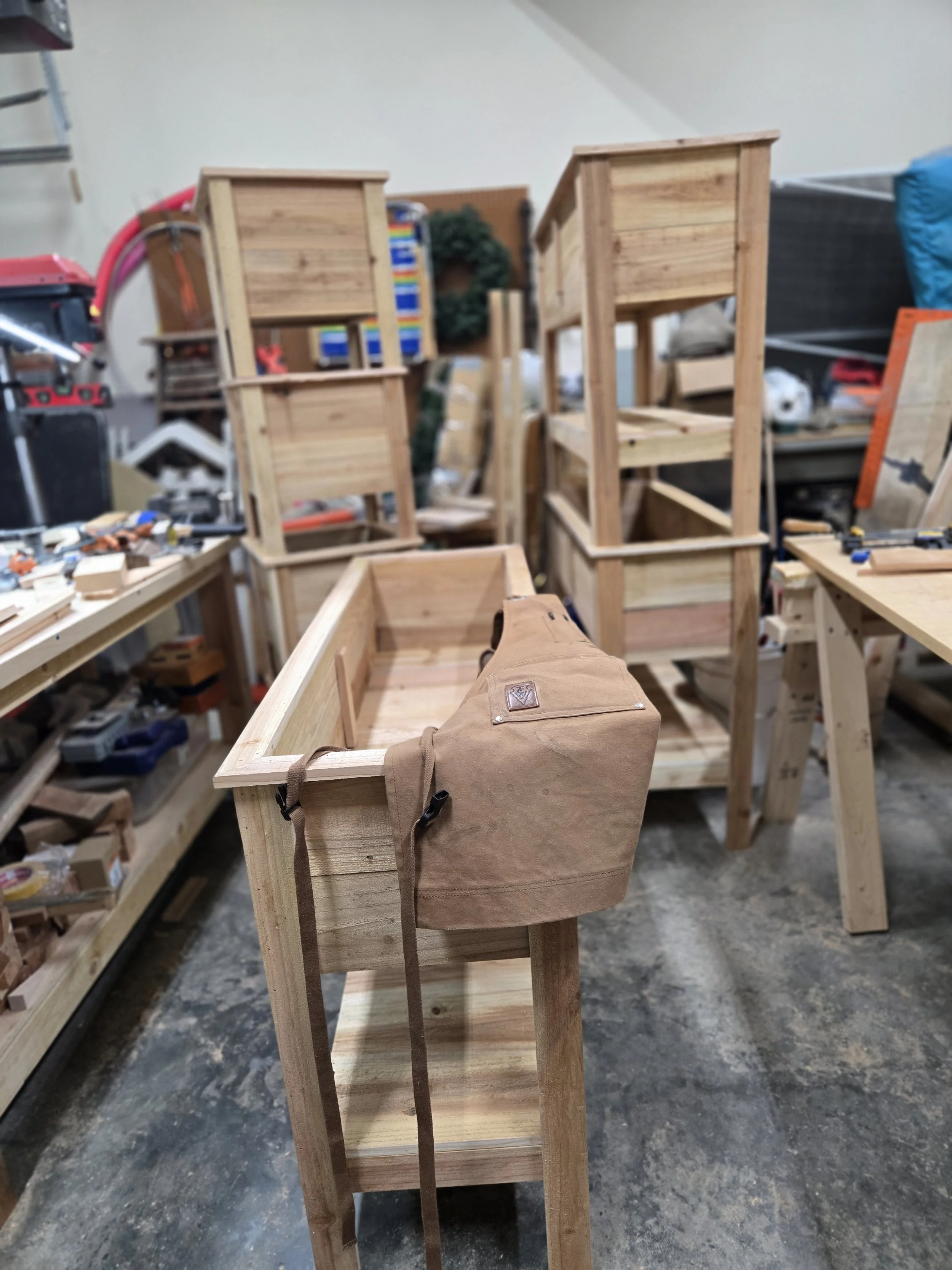 A woodworking workshop showing wooden furniture in progress, including multiple wooden shelves and a workbench with tools and a brown shop apron.